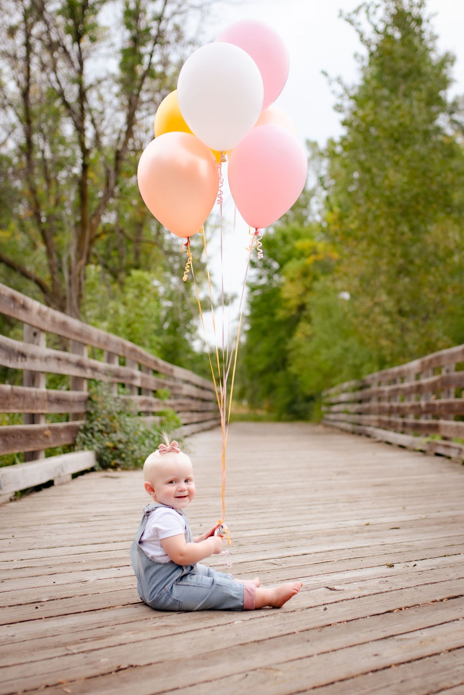 Baby in overalls sits on wooden walkway holding colorful balloons with string decorations.