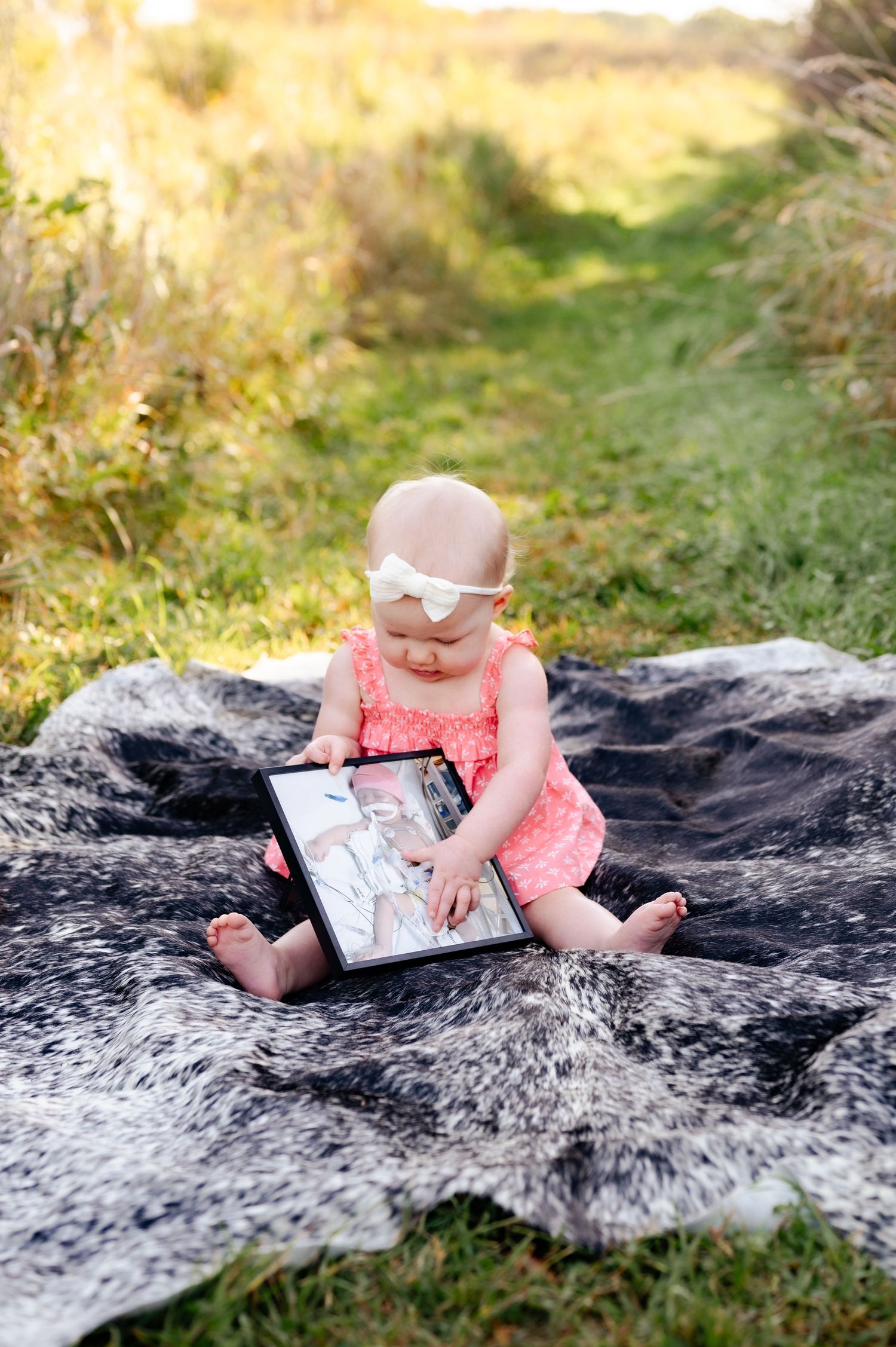 Baby in coral dress sits on fuzzy blanket looking at photo on digital tablet during golden hour outdoor photoshoot.