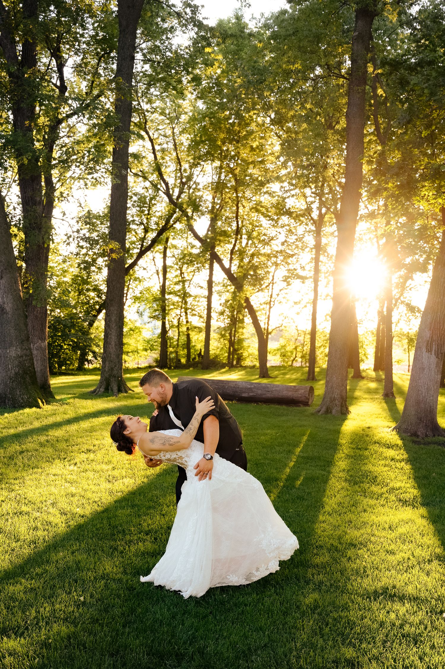 Romantic sunset dip kiss between newlyweds on lush green lawn with sunlit trees.