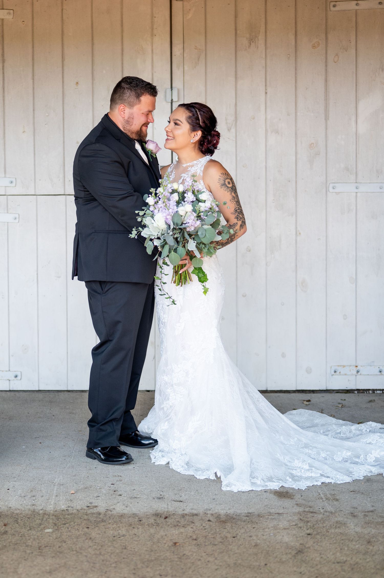 Bride holding purple and white bouquet poses with groom against rustic barn backdrop.