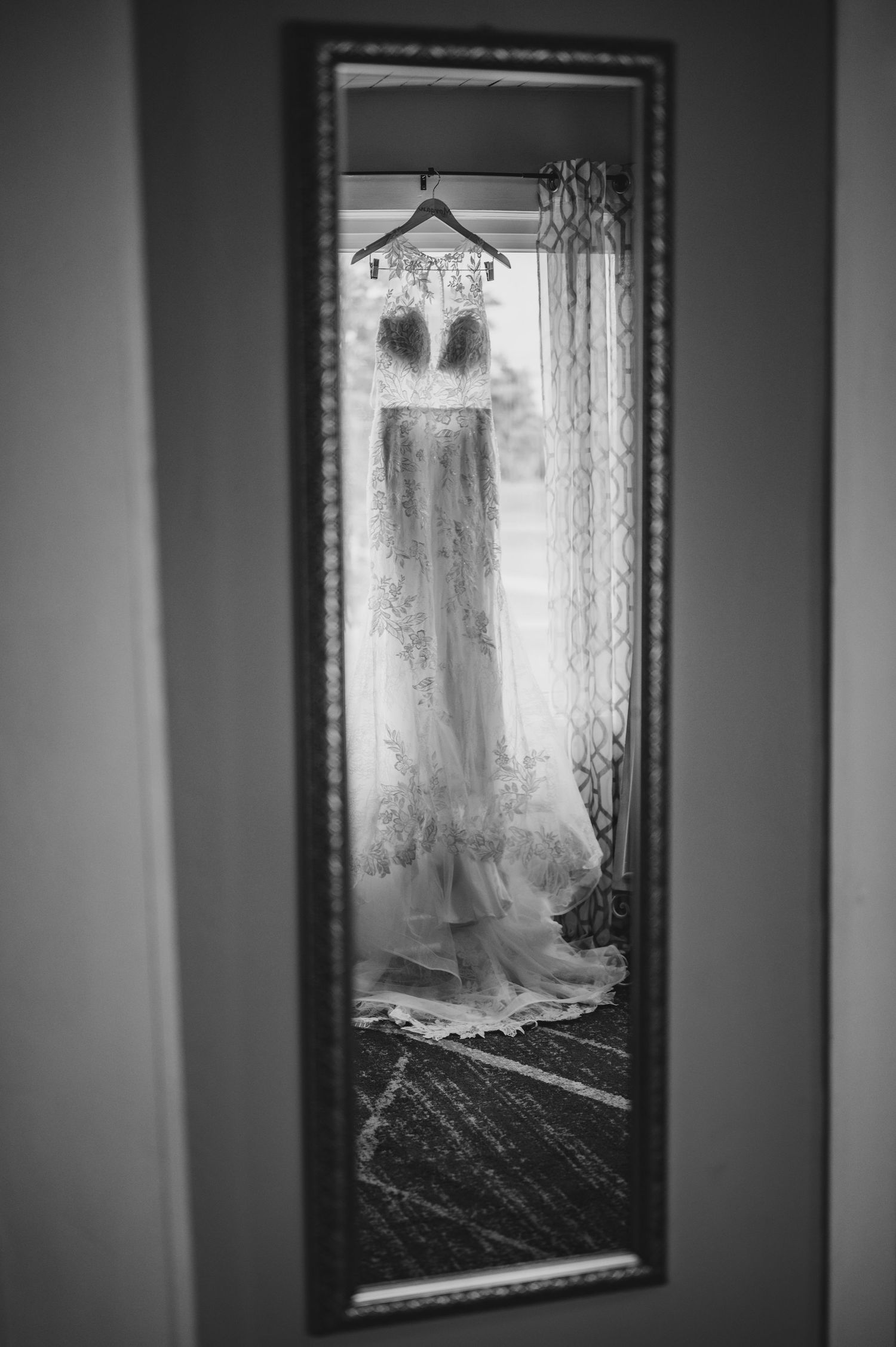 A wedding dress hanging in an ornate mirror reflection creating an ethereal black and white image.