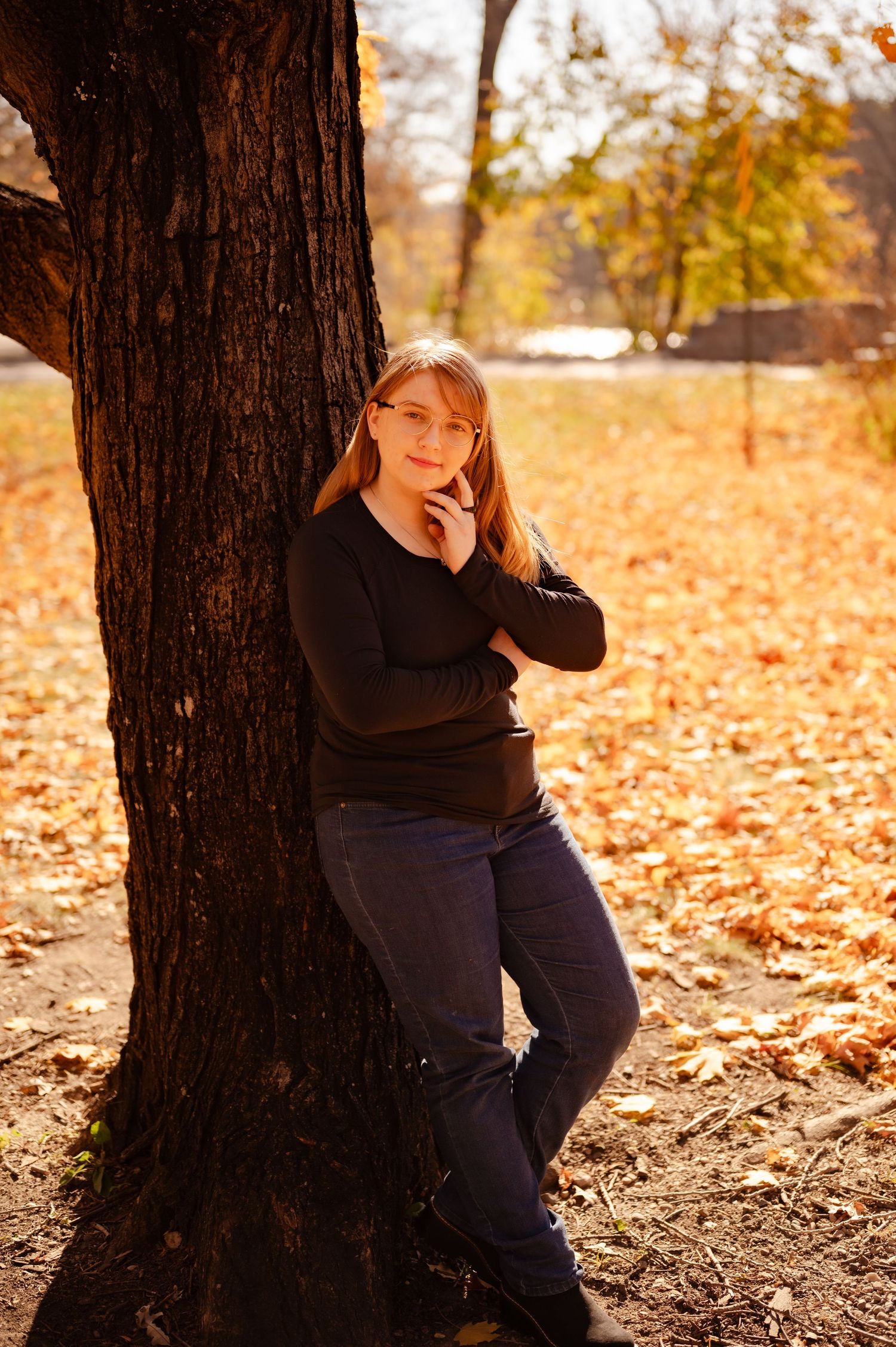 Figure in dark clothing sits thoughtfully against large tree trunk surrounded by fallen autumn leaves.