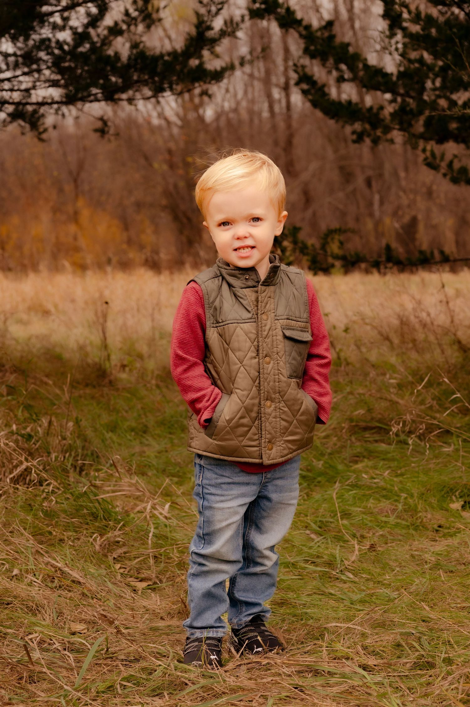 A young child stands in a grassy field wearing a brown quilted vest, red shirt and jeans on an autumn day.