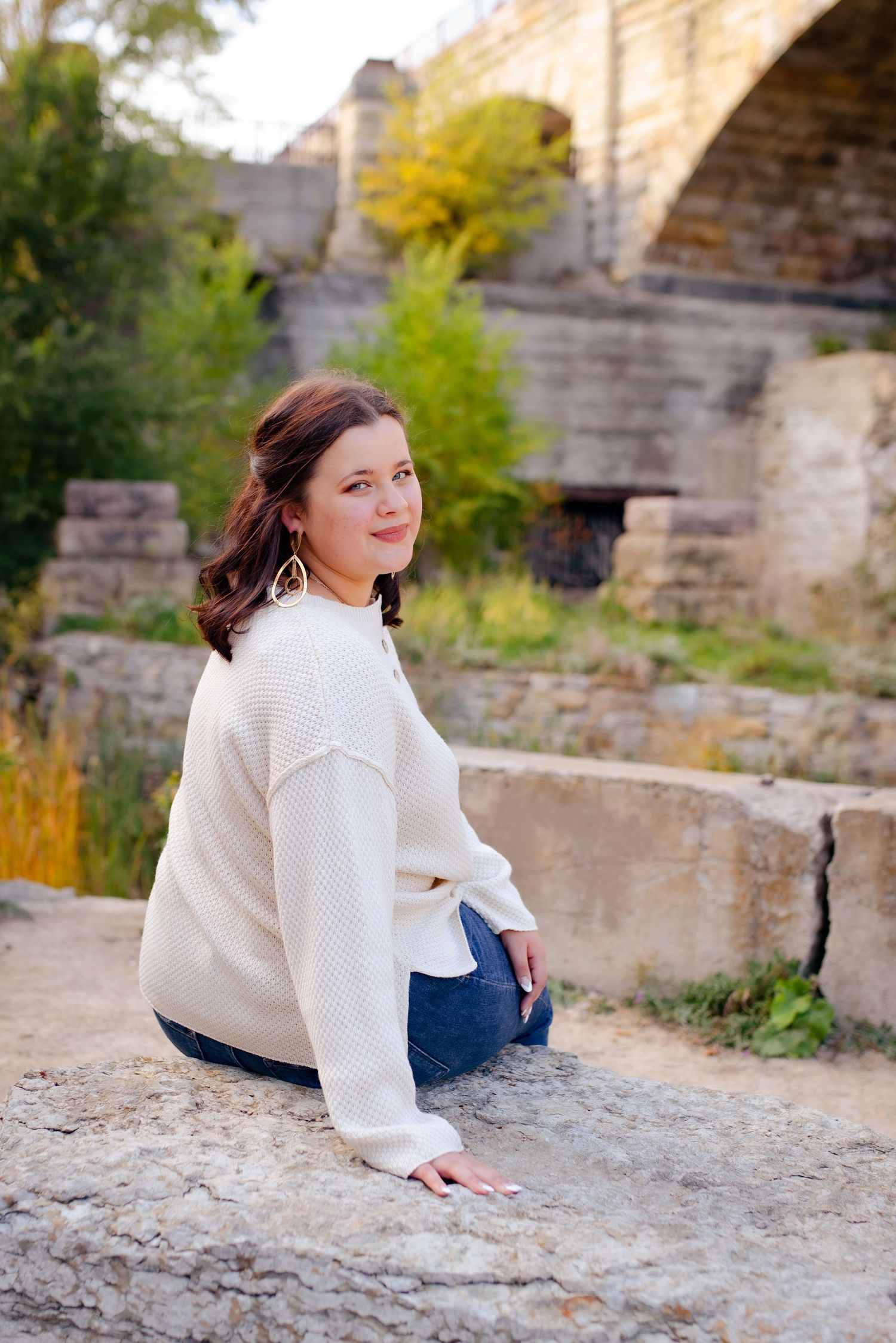 Person in cream sweater sits on stone ledge near bridge with fall colors in background.