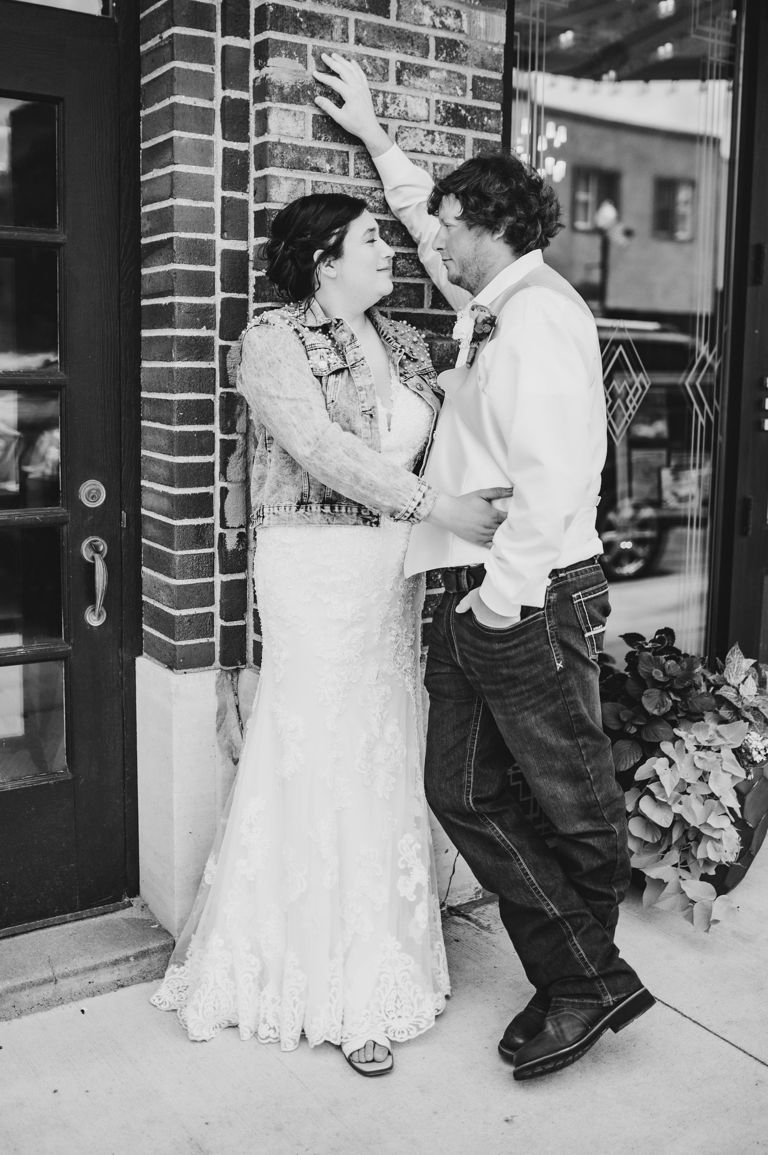 A playful moment between a bride in a lace dress and groom in casual attire against a brick wall exterior.
