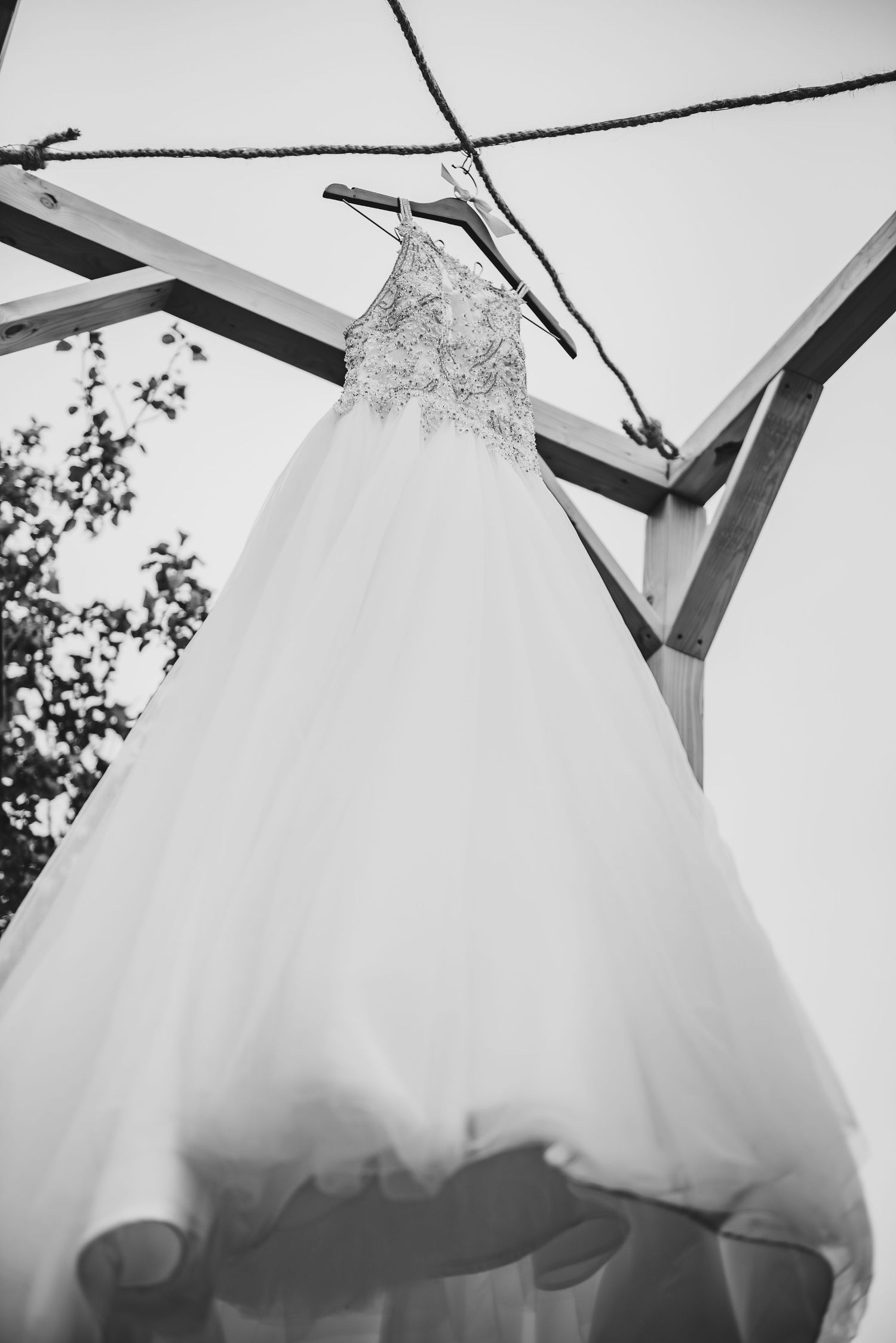 Black and white photo of a wedding dress hanging from wooden beams against sky.