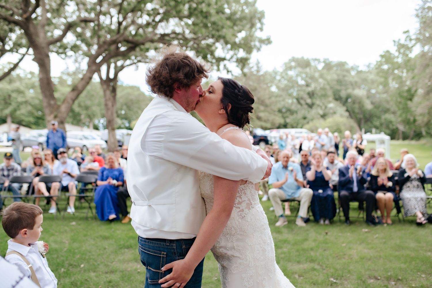 Romantic celebration kiss at outdoor wedding with guests watching in background.