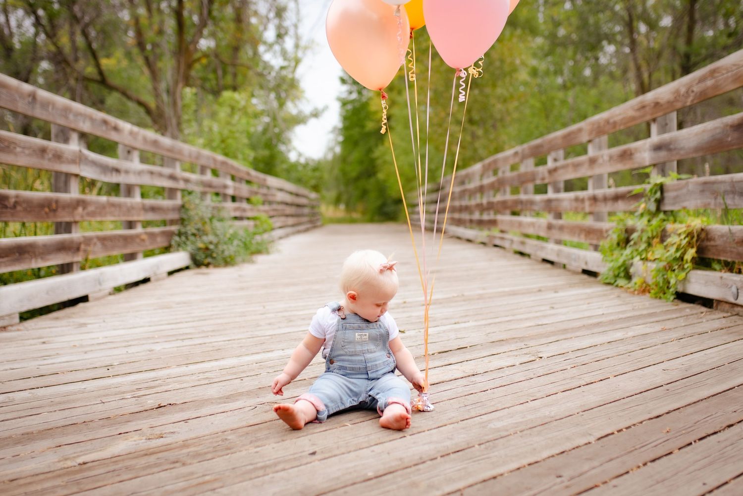 Baby in overalls sits on boardwalk holding pink and peach balloons with trees in background.
