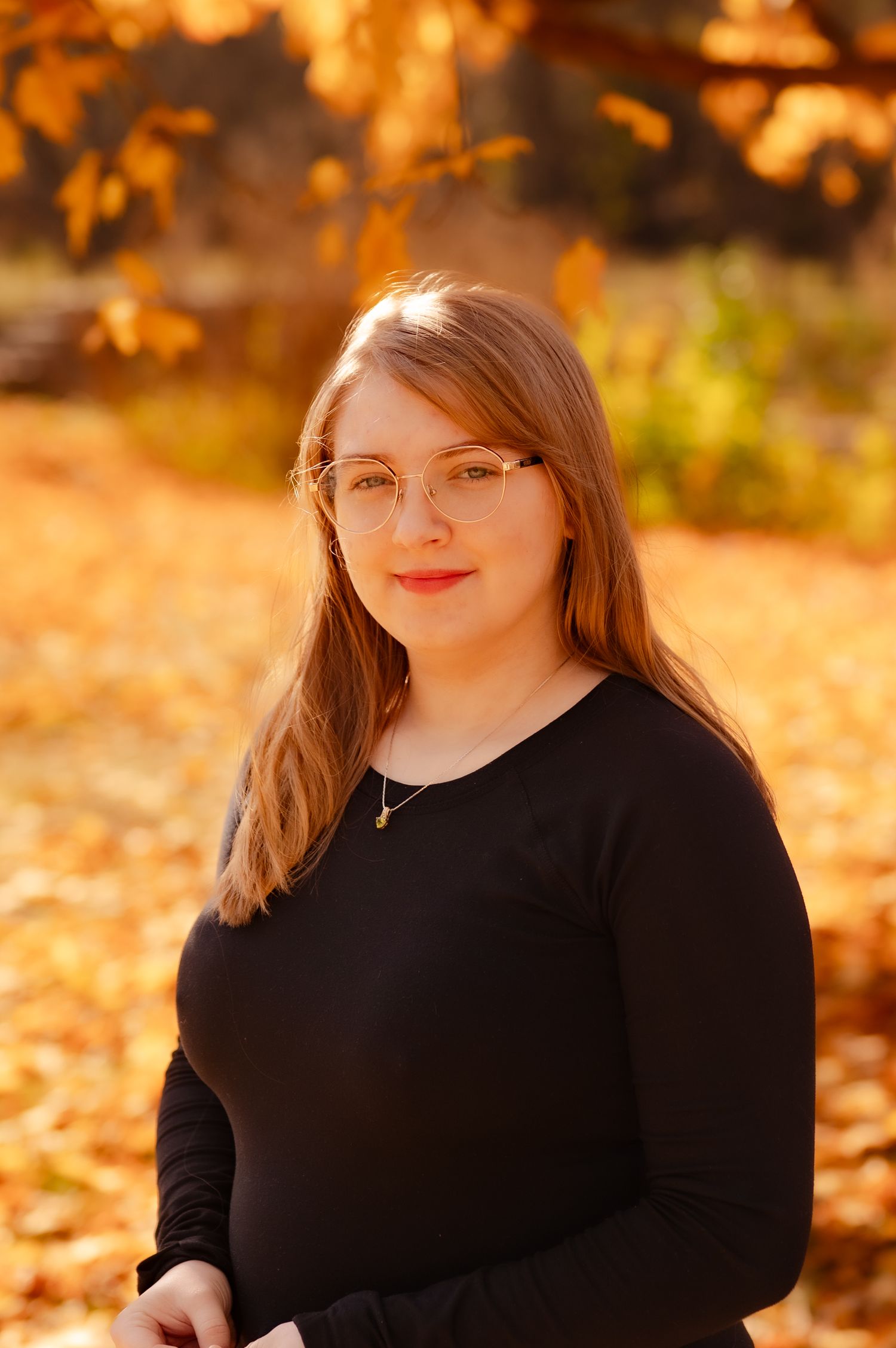 Person in black sweater poses against warm autumn backdrop with golden sunlit foliage.