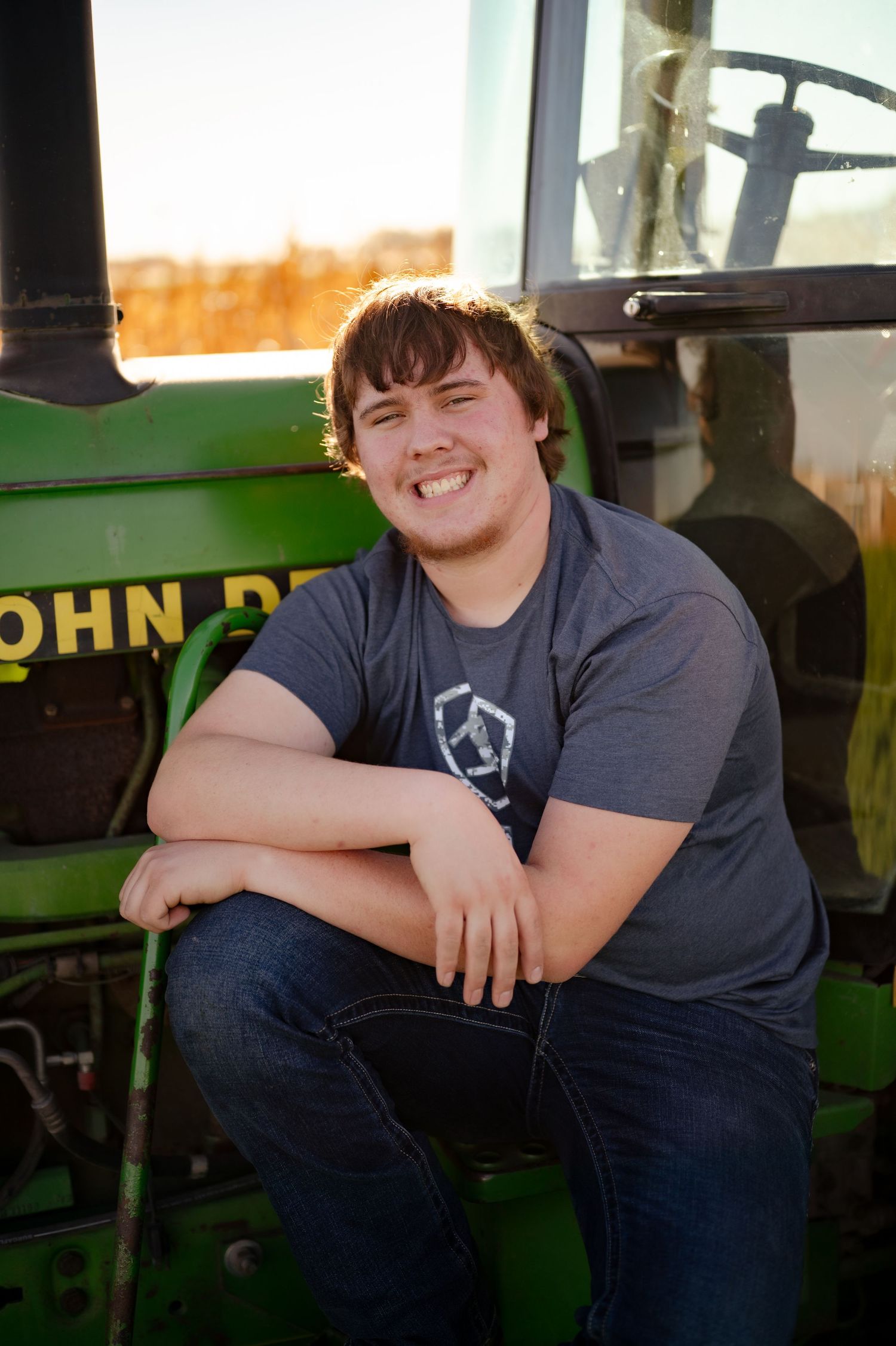 Someone in a gray t-shirt sits smiling on a green John Deere tractor during a sunny outdoor photo session.
