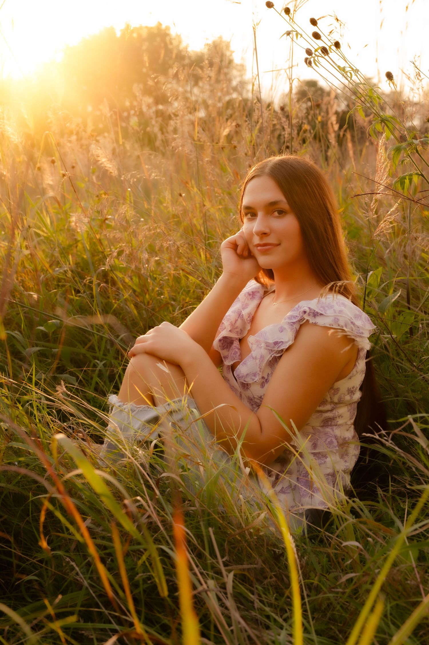 A woman in a floral dress relaxes in tall grass during golden hour sunset.