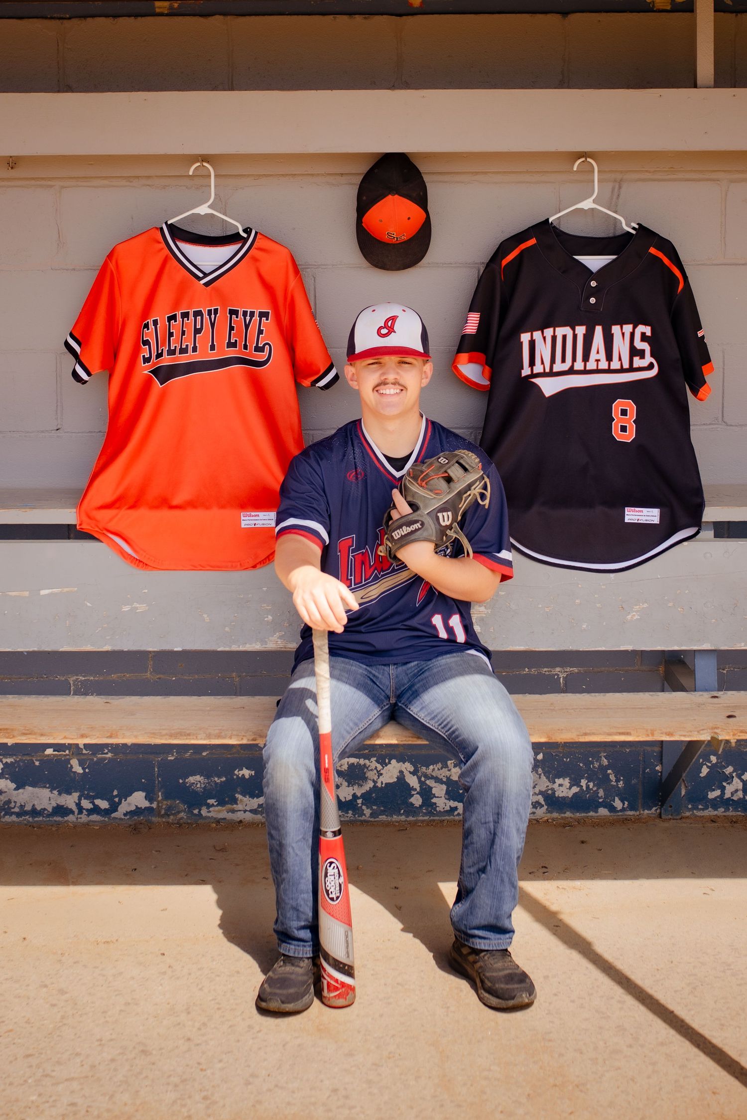 Baseball player sits in dugout with team jerseys hanging behind on wall.