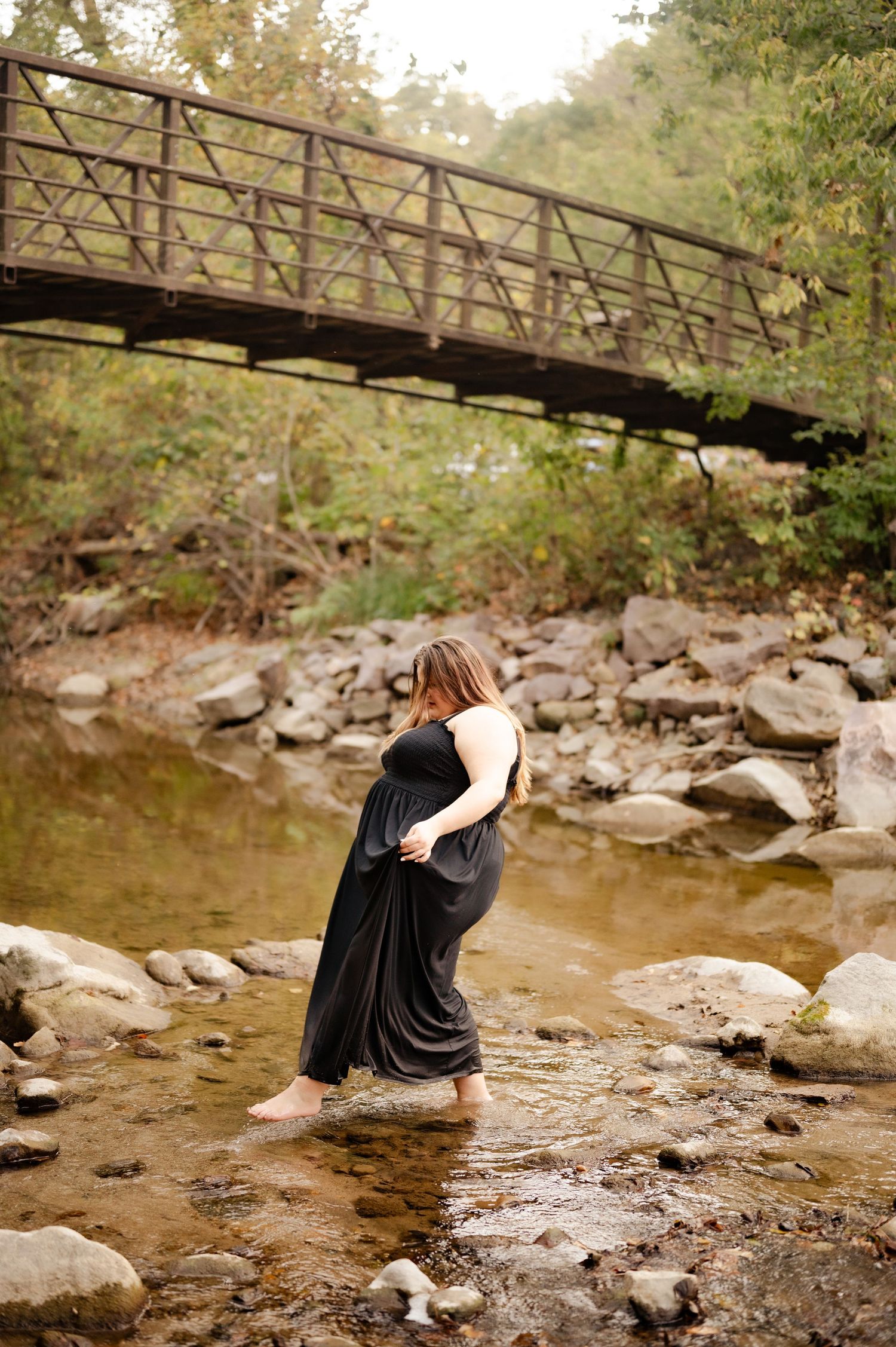 Person in black dress standing in river water beneath rustic bridge.