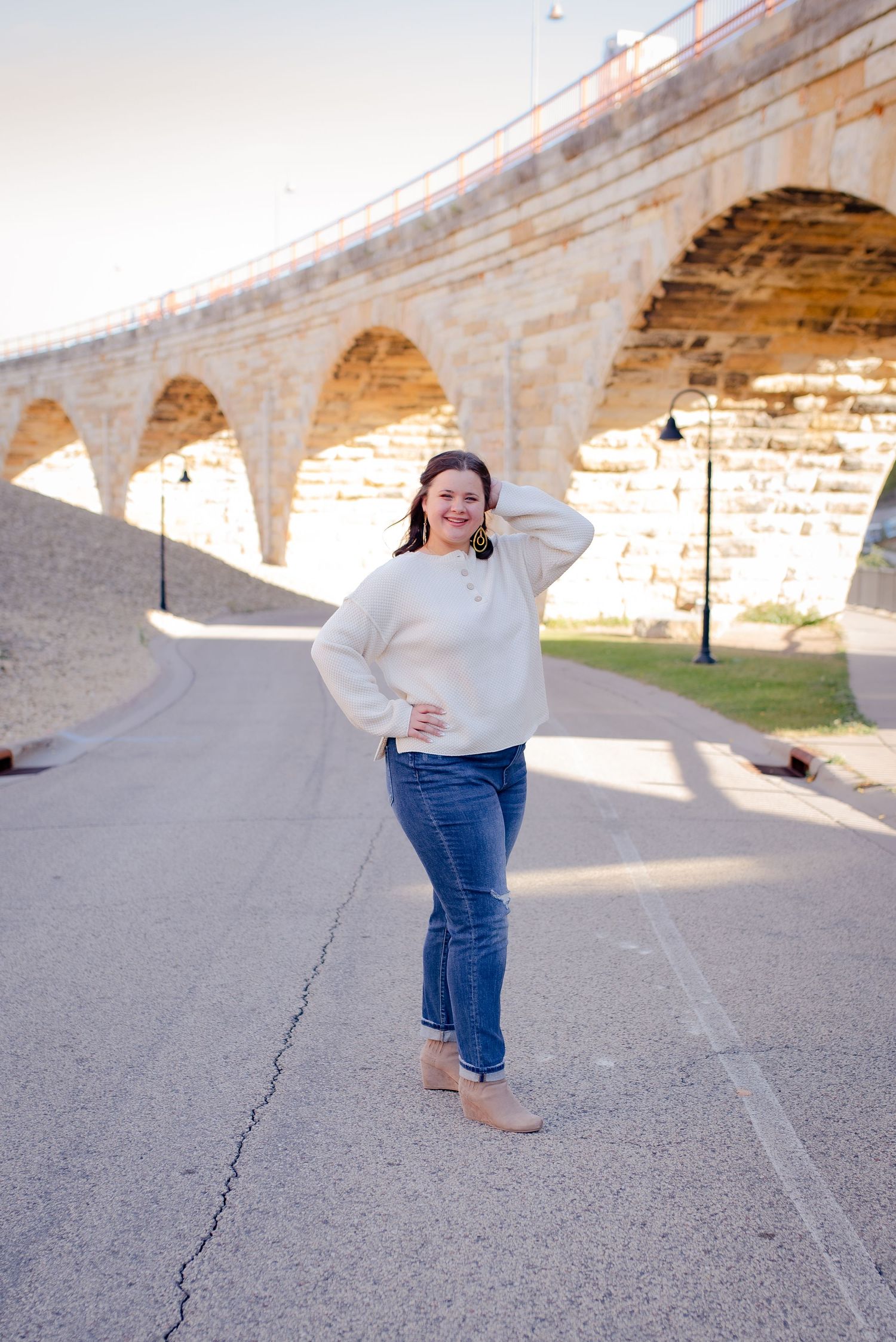 Someone poses in a cream sweater and jeans beneath a stone arch bridge on a sunny day.