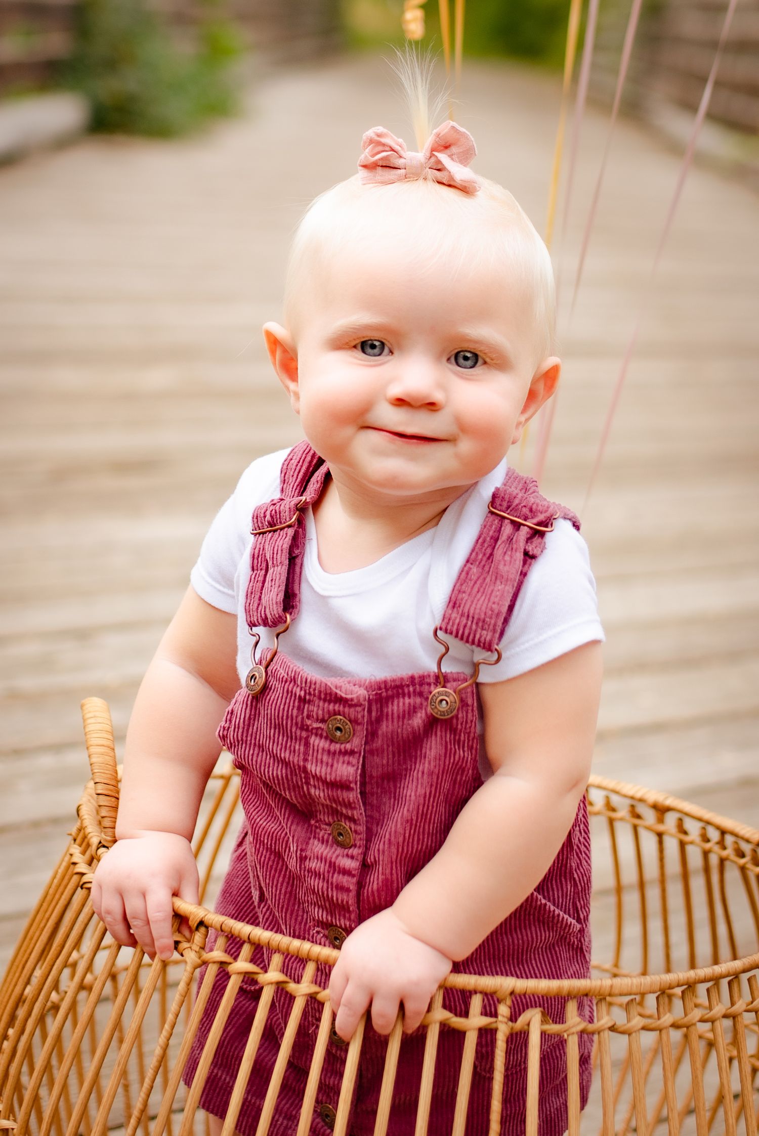 Baby wearing corduroy overalls sits in wicker basket with pink bow accessory.