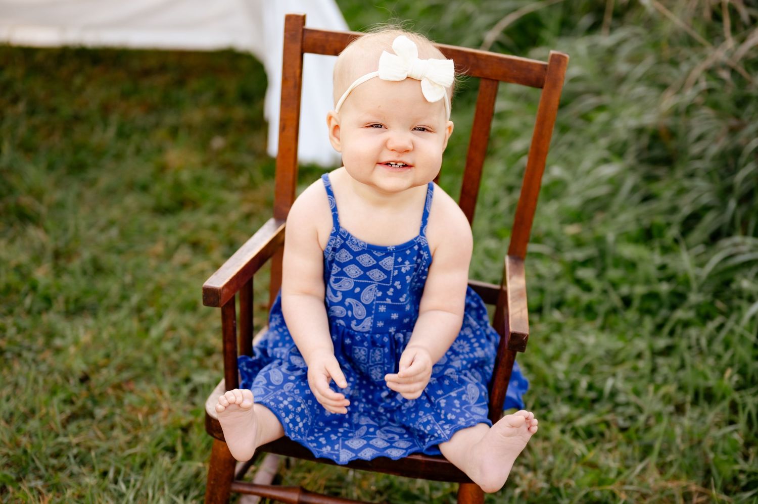 A cheerful toddler in a blue floral dress sits in a wooden chair outdoors.