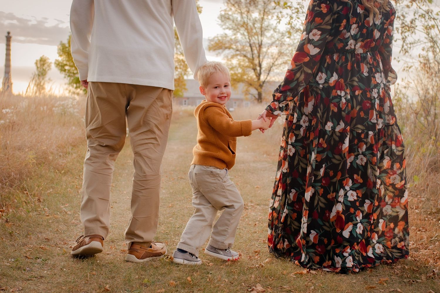 A small child in orange holds hands with adults wearing floral dress and khaki pants outdoors.