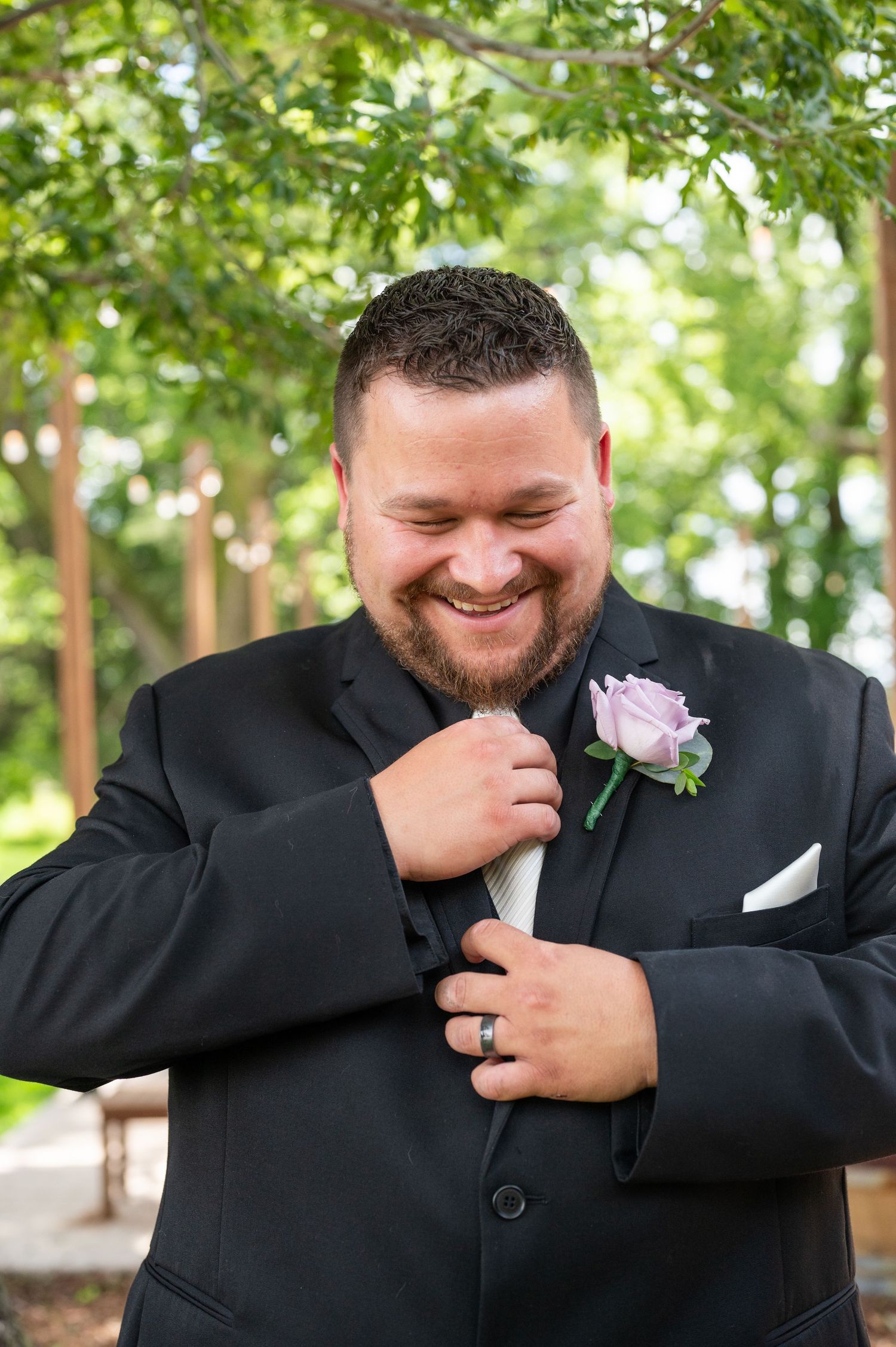 Happy groom wearing black suit and pink boutonniere smiles while adjusting his tie.