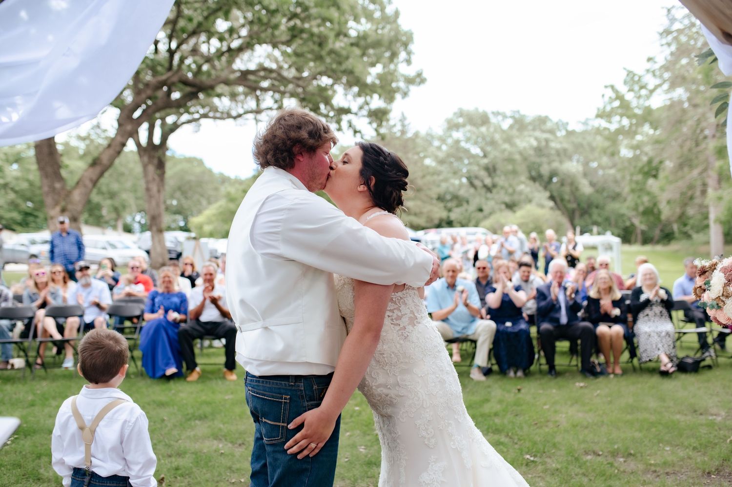Couple shares their first kiss at outdoor wedding with guests in background.