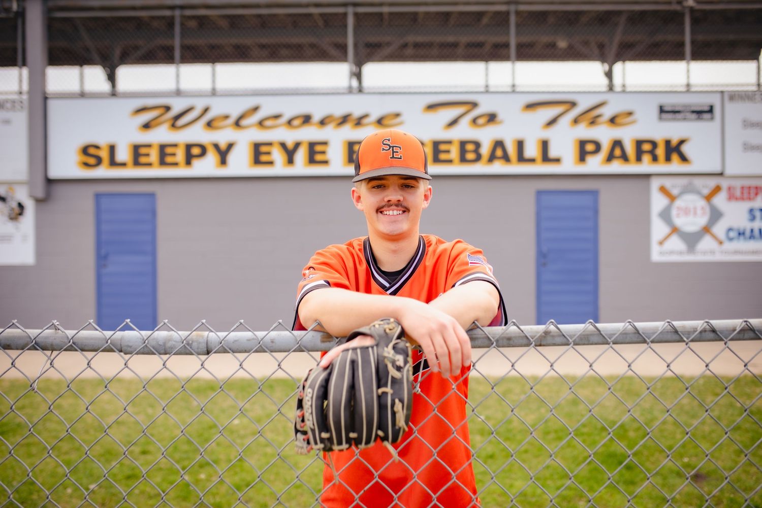 Baseball player in orange uniform stands at Sleepy Eye Baseball Park holding glove.