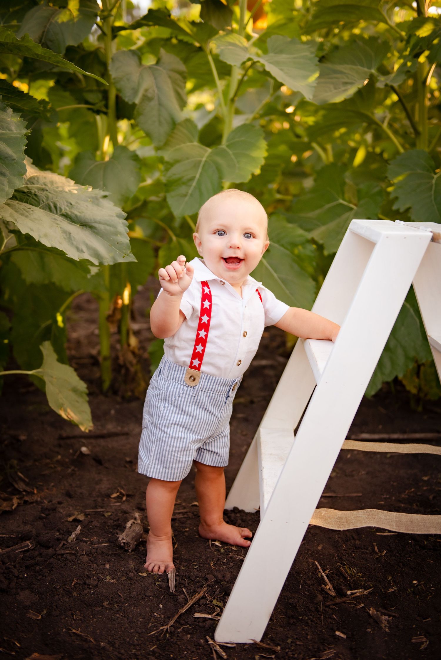 Baby in striped overalls standing by white ladder against green garden background.