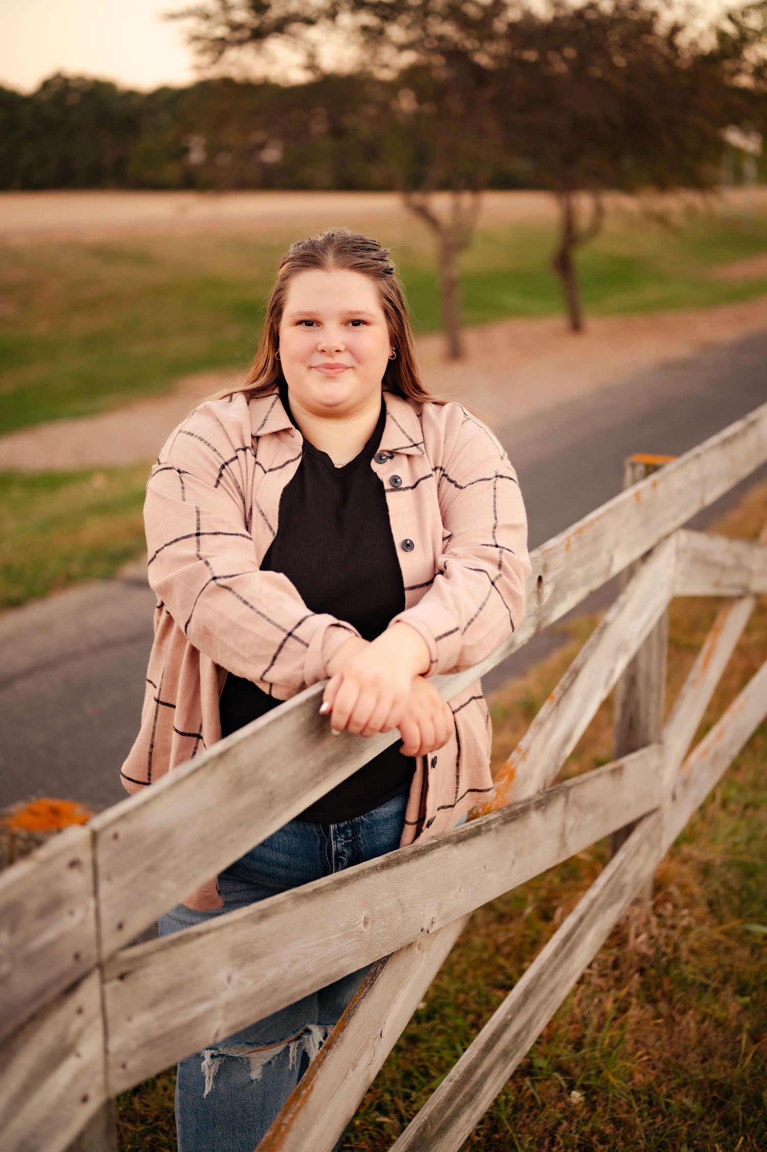 Person leaning on wooden fence wearing beige jacket and jeans during sunset.