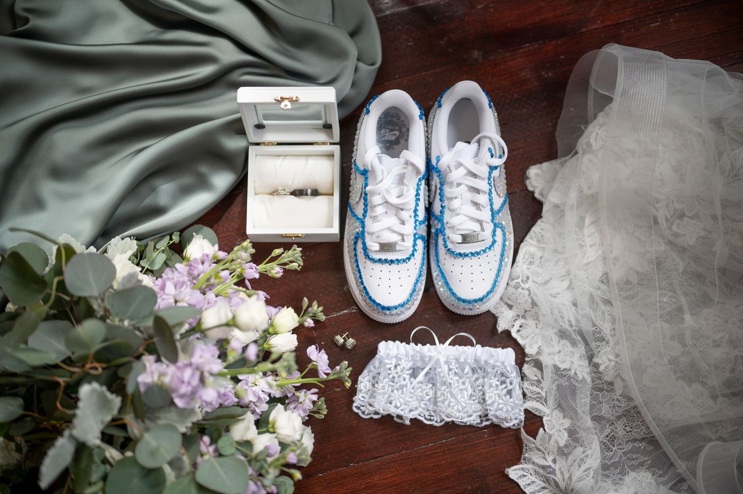 Wedding accessories including blue sneakers, garter, flowers and jewelry box arranged on wooden surface.