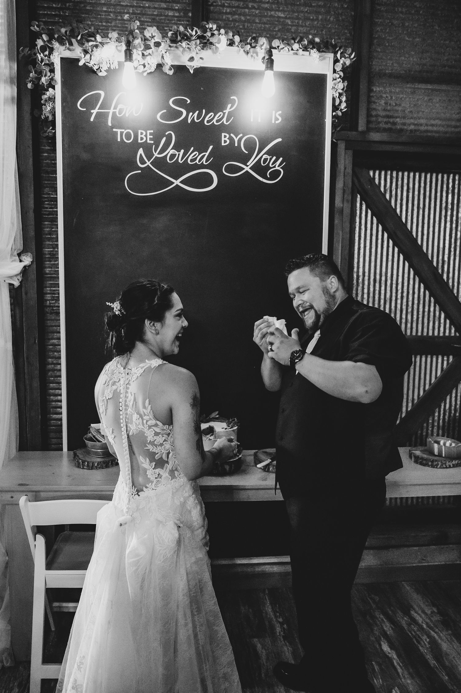 Black and white photo of a couple laughing together at a wedding reception with a decorated chalkboard sign above.