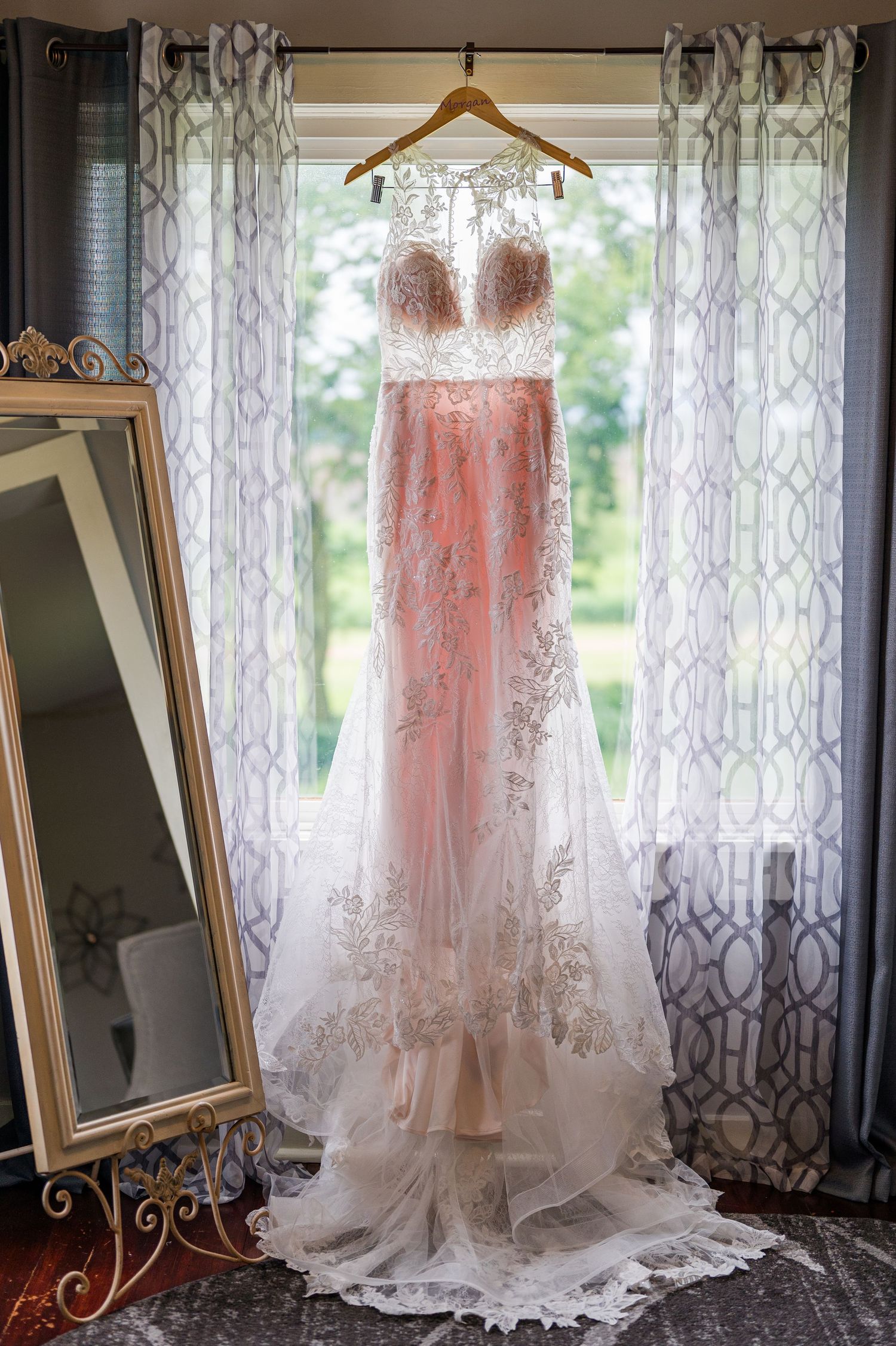 Blush lace wedding dress hanging in window with flowing train and delicate details.