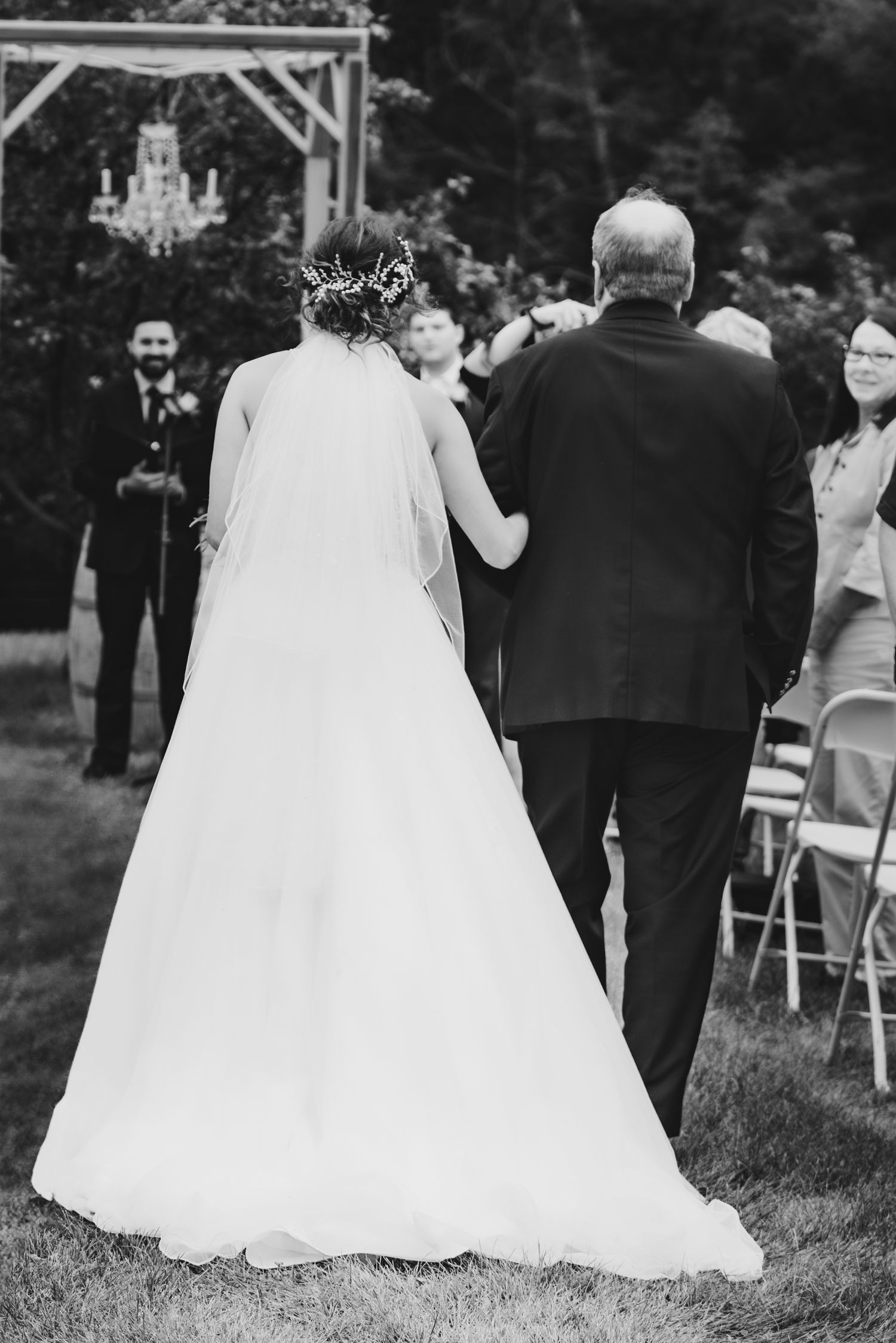 A bride in a flowing white gown walks down the aisle during an outdoor garden wedding ceremony with a chandelier overhead.