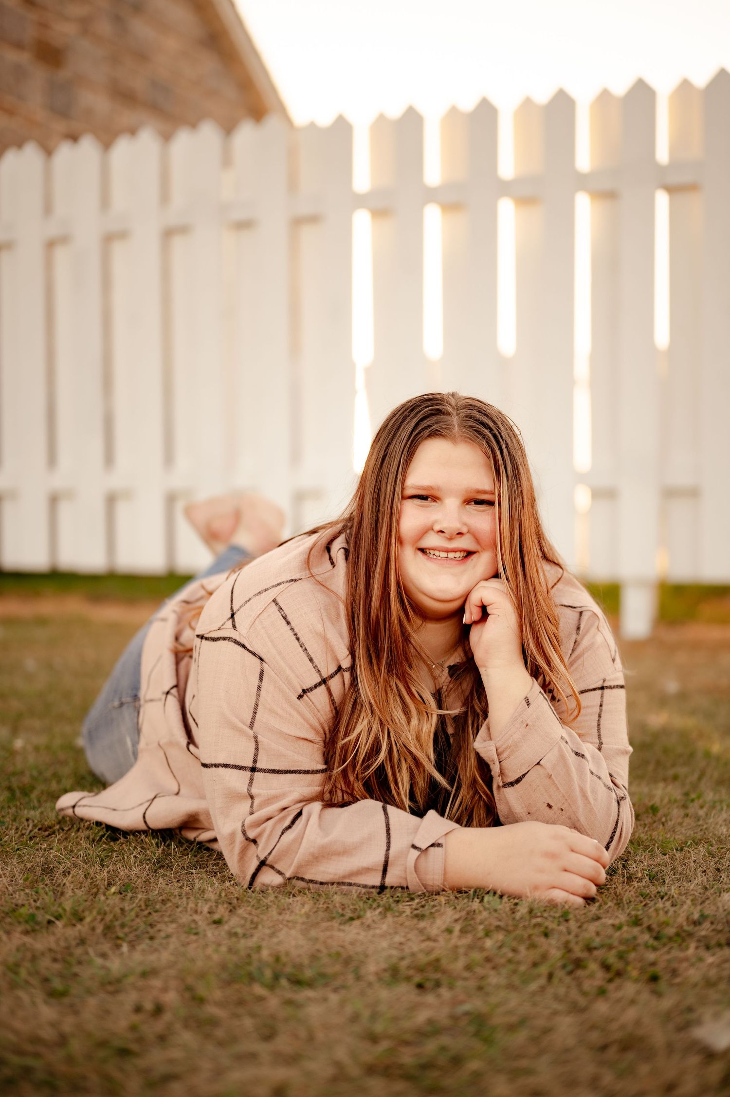 Person lying on grass wearing tan jacket with white picket fence in background during golden hour.