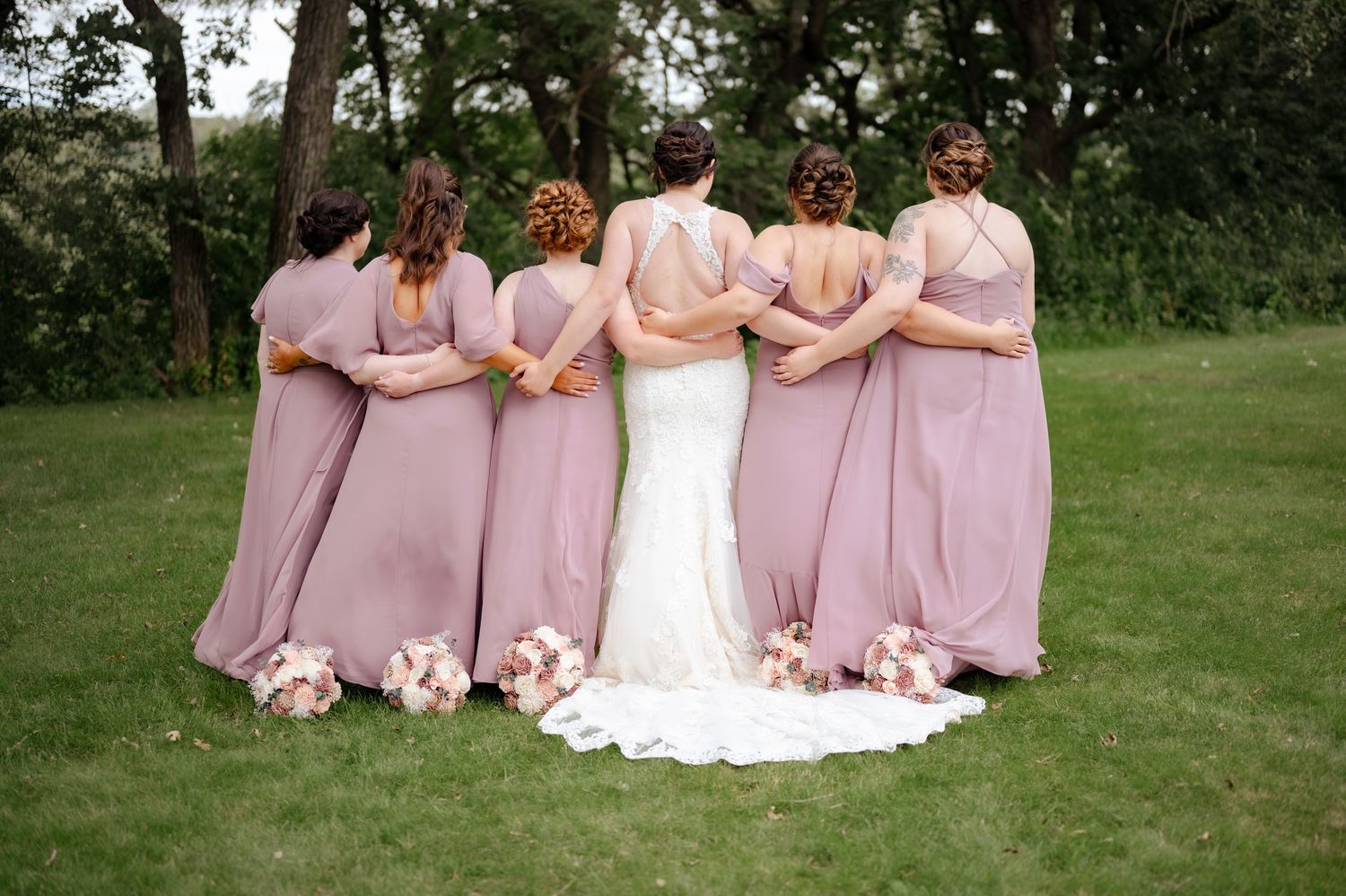 Group wearing matching pink bridesmaid dresses standing with bride in white gown outdoors.