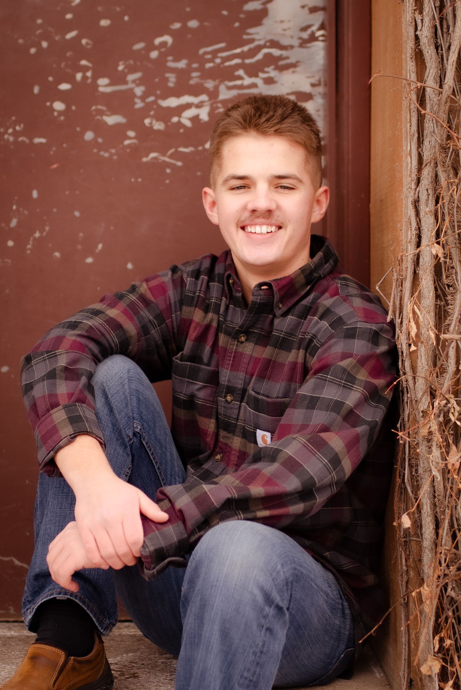 Casual portrait sitting by weathered wall wearing plaid shirt and denim.