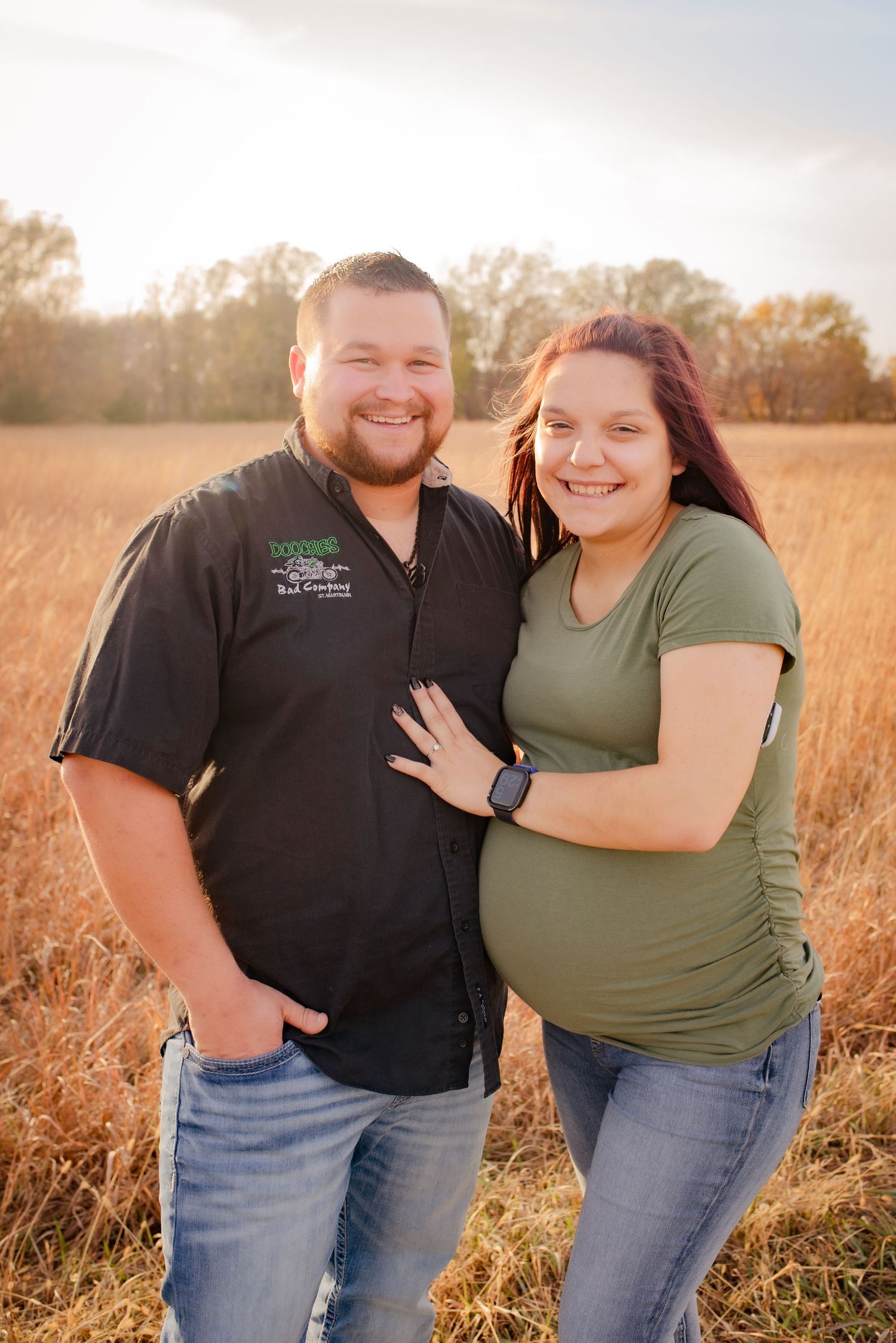 Expecting couple poses together in autumn field wearing casual attire.