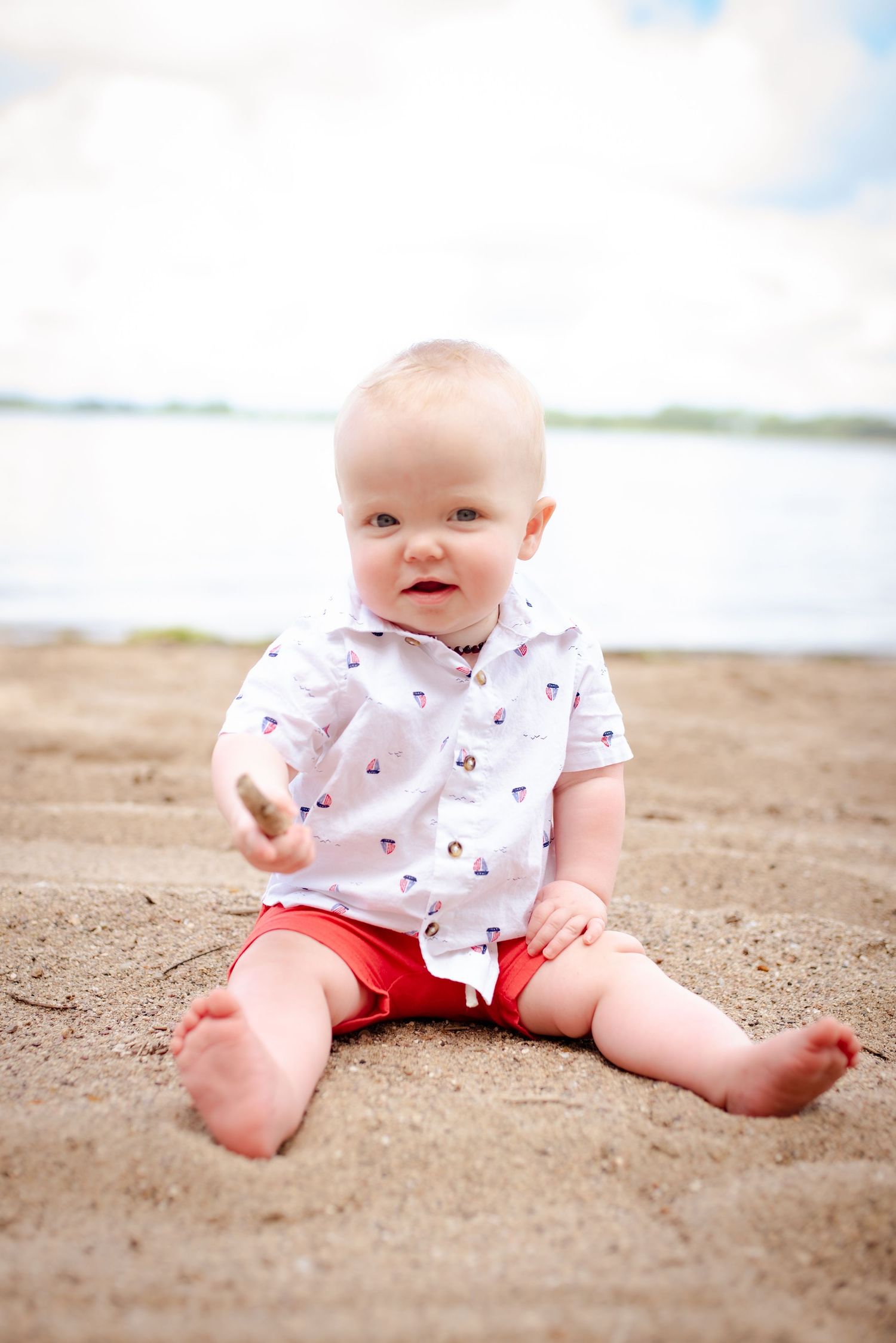 A baby wearing a patterned shirt and coral shorts sits on a sandy beach by the water.