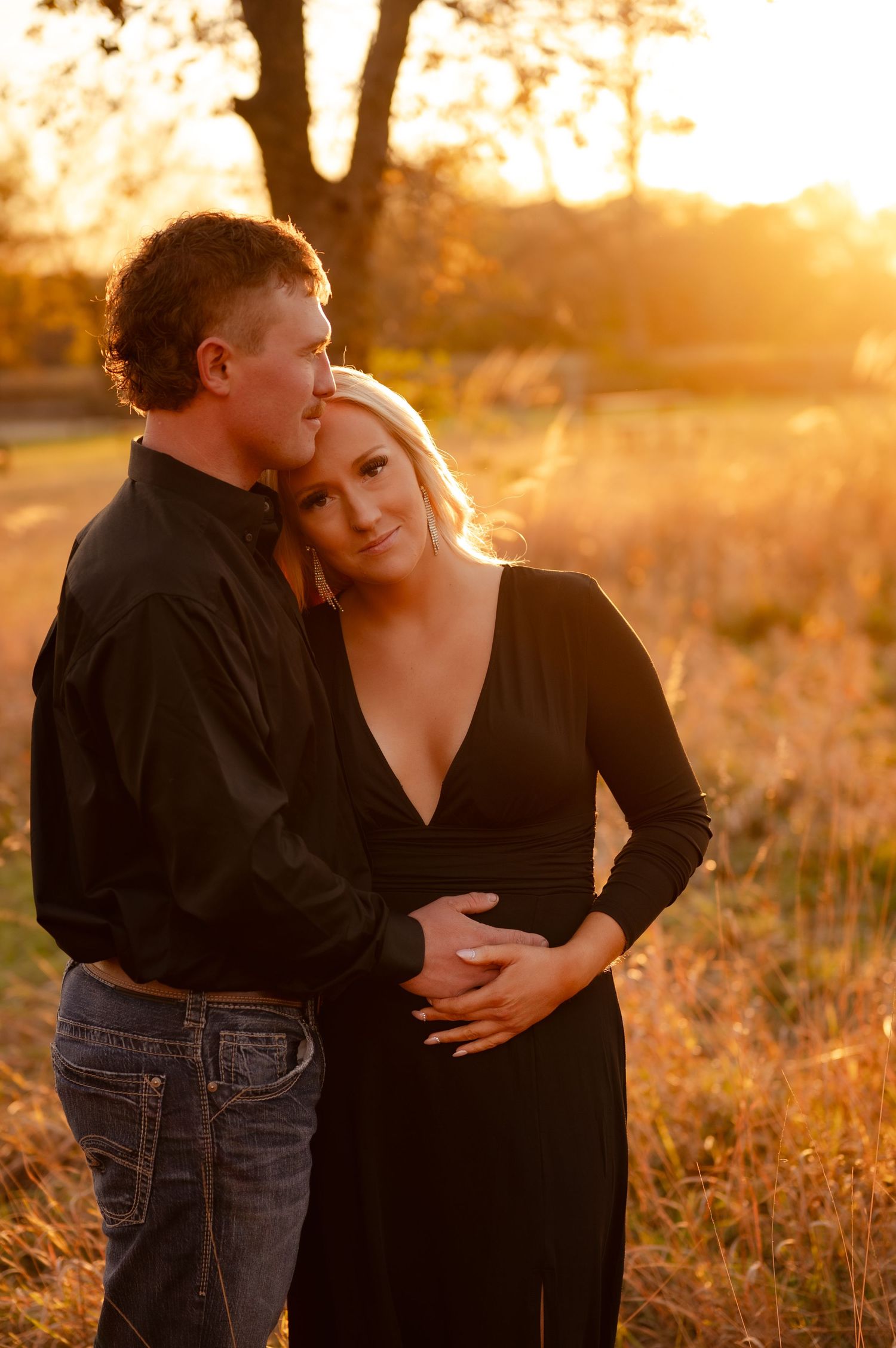 A romantic maternity photoshoot at golden hour in a field with warm sunset lighting and tall grass.