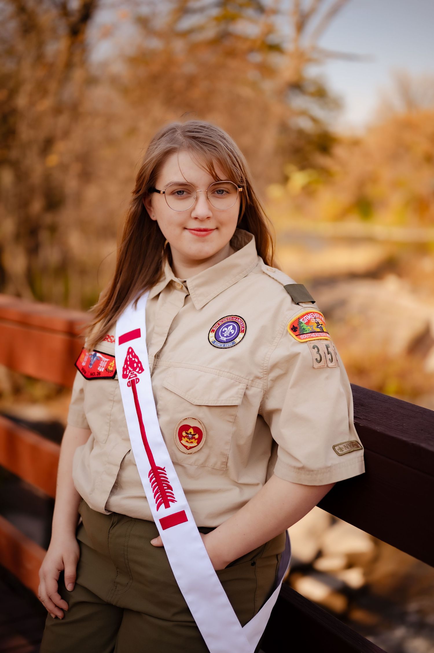 Scout member wearing a tan uniform with badges and white sash poses outdoors during fall season.