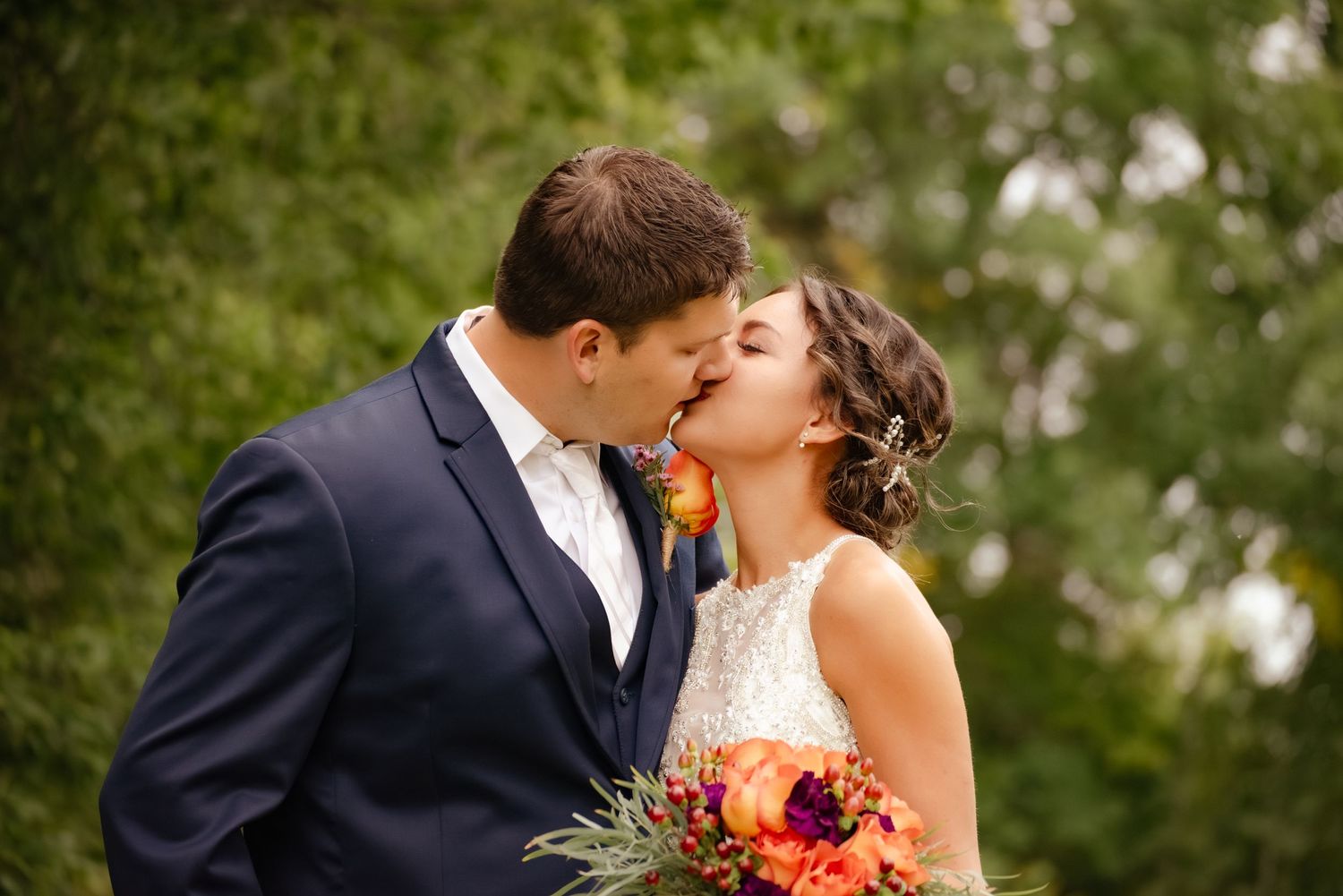 Romantic wedding couple shares a kiss while holding a vibrant autumn bouquet in a garden setting.