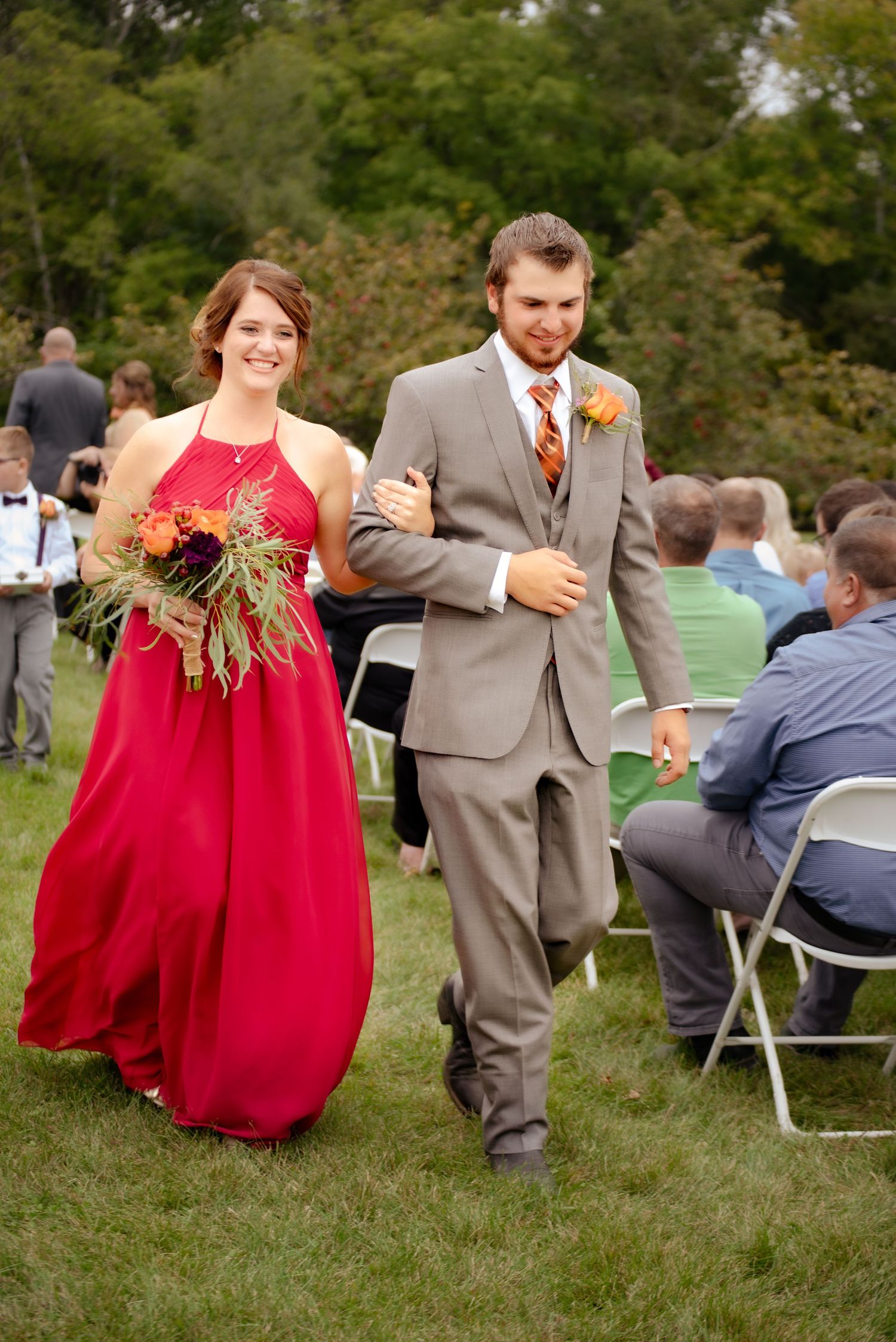A couple walks down the aisle at an outdoor wedding ceremony with red floral decorations.