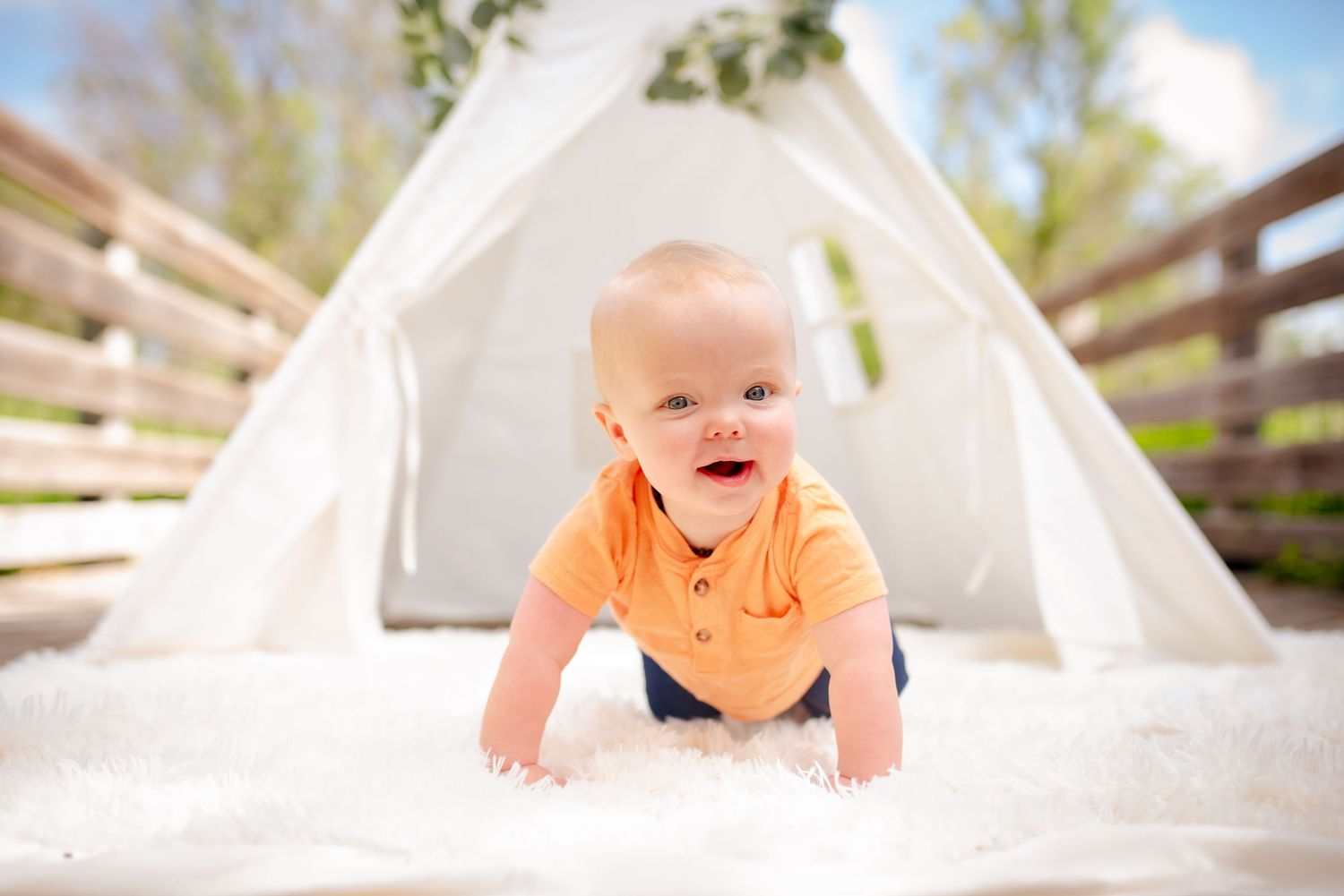 A smiling baby crawls inside a white teepee tent outdoors.