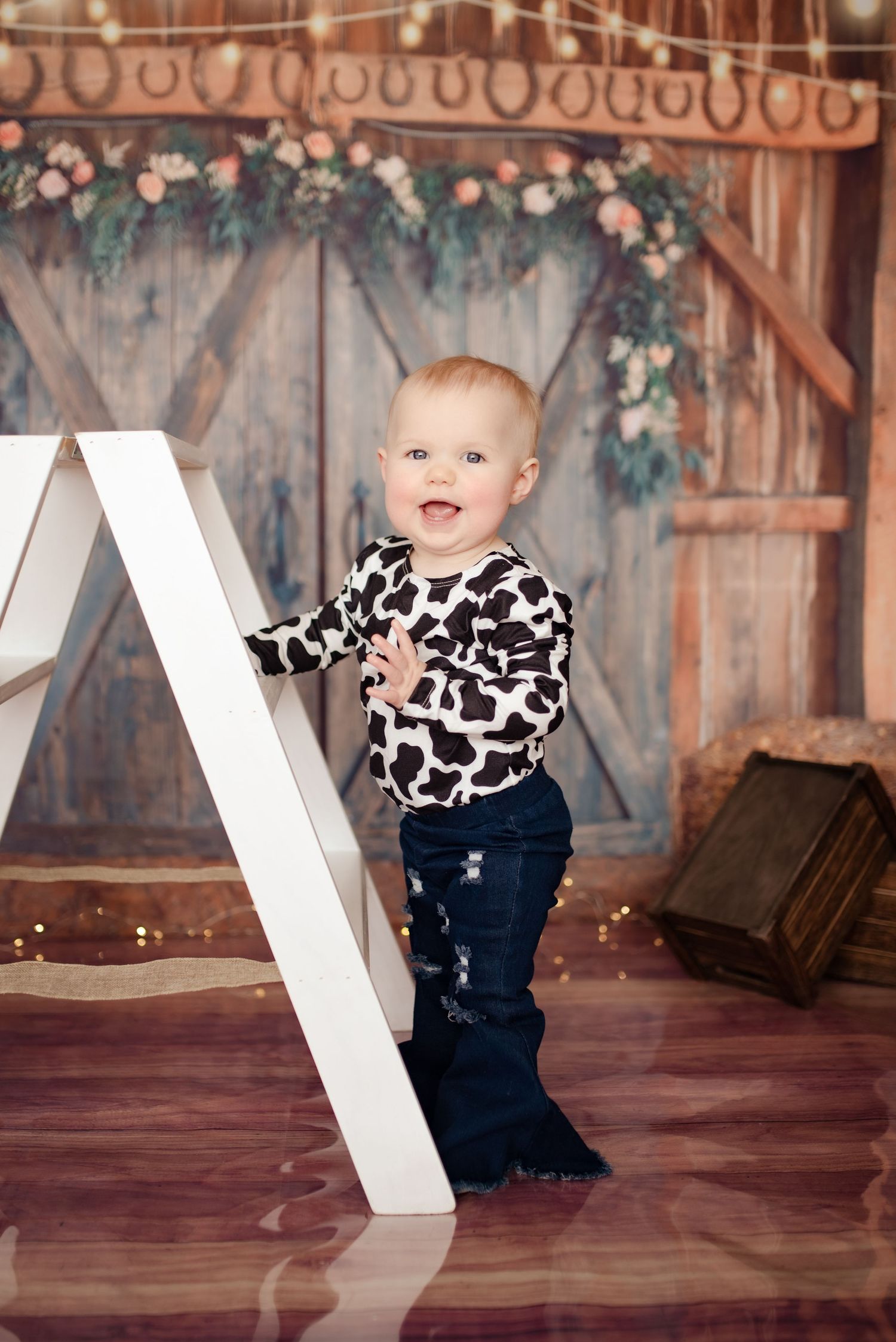 Toddler in cow print top standing by white ladder against rustic barn backdrop with floral garland.