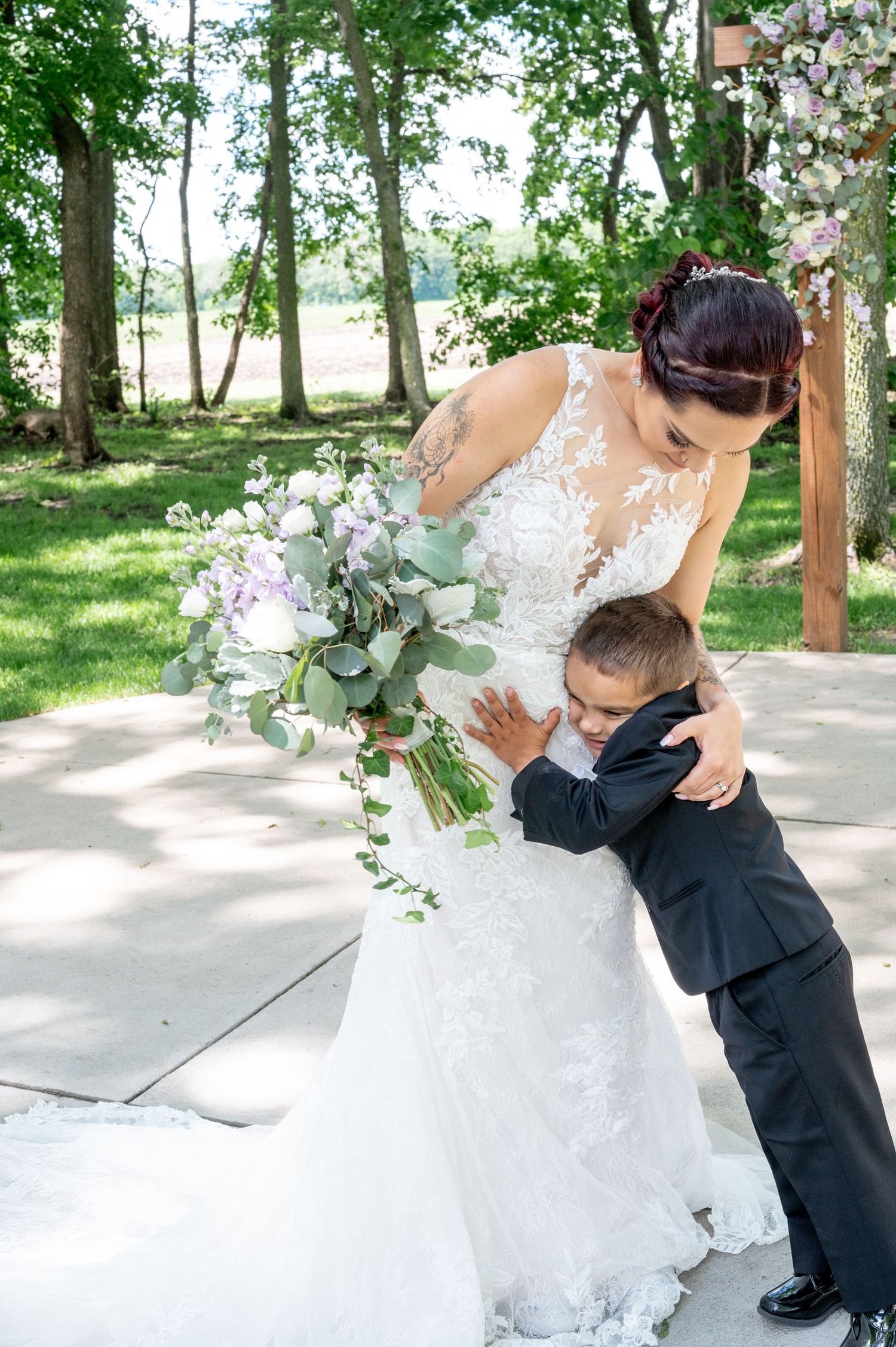 A tender embrace between a young attendee and bride in a lace wedding dress with a cascading bouquet in an outdoor setting.