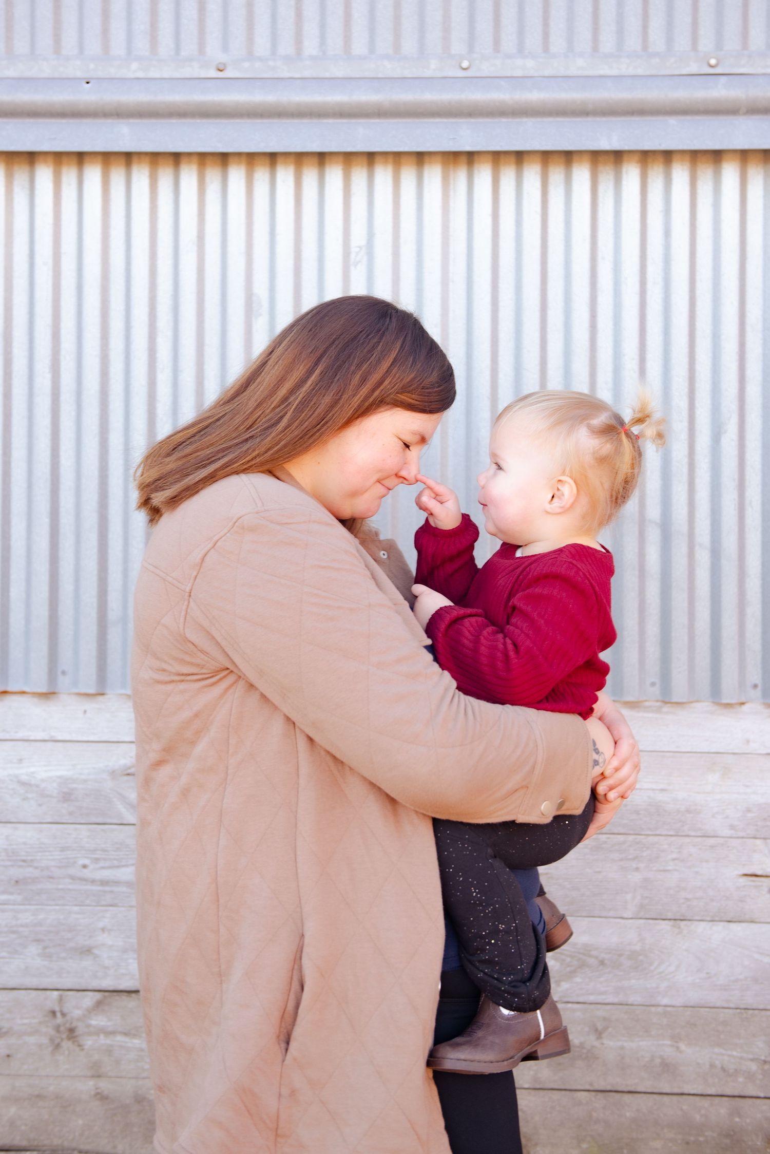 Mother in tan coat holding and gazing lovingly at baby in red sweater against corrugated wall.