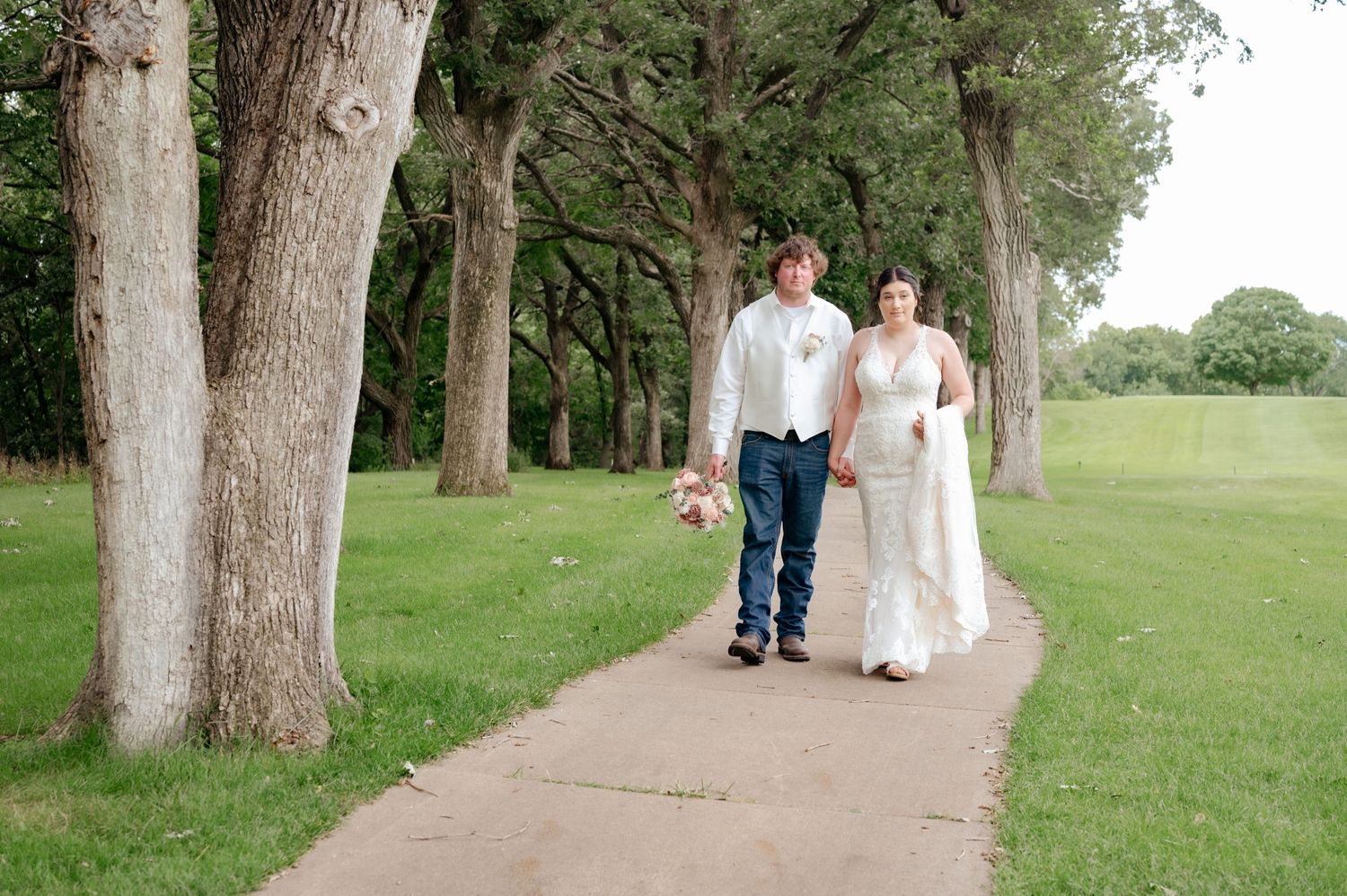 Newlyweds walking together on concrete path lined with tall trees.