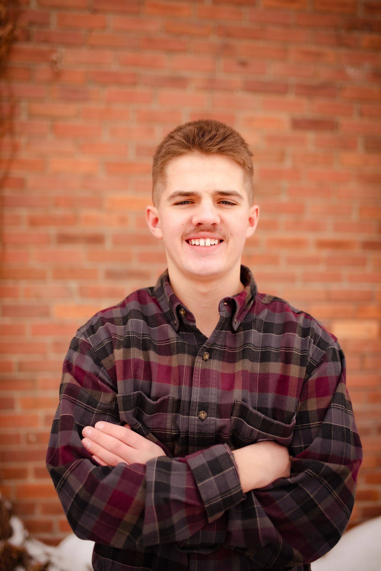 Portrait against brick wall with arms crossed wearing plaid flannel shirt.
