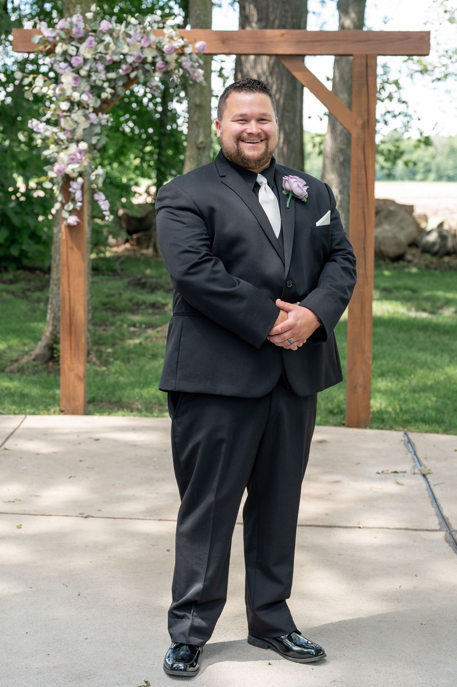 Wedding party member in black tuxedo standing in front of wooden arch with flowers.