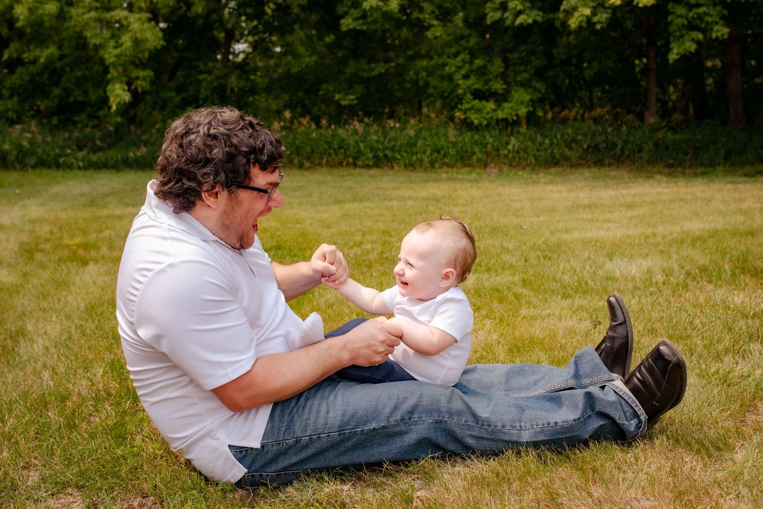 A playful moment between parent and baby sitting on grass during a sunny day outdoors.