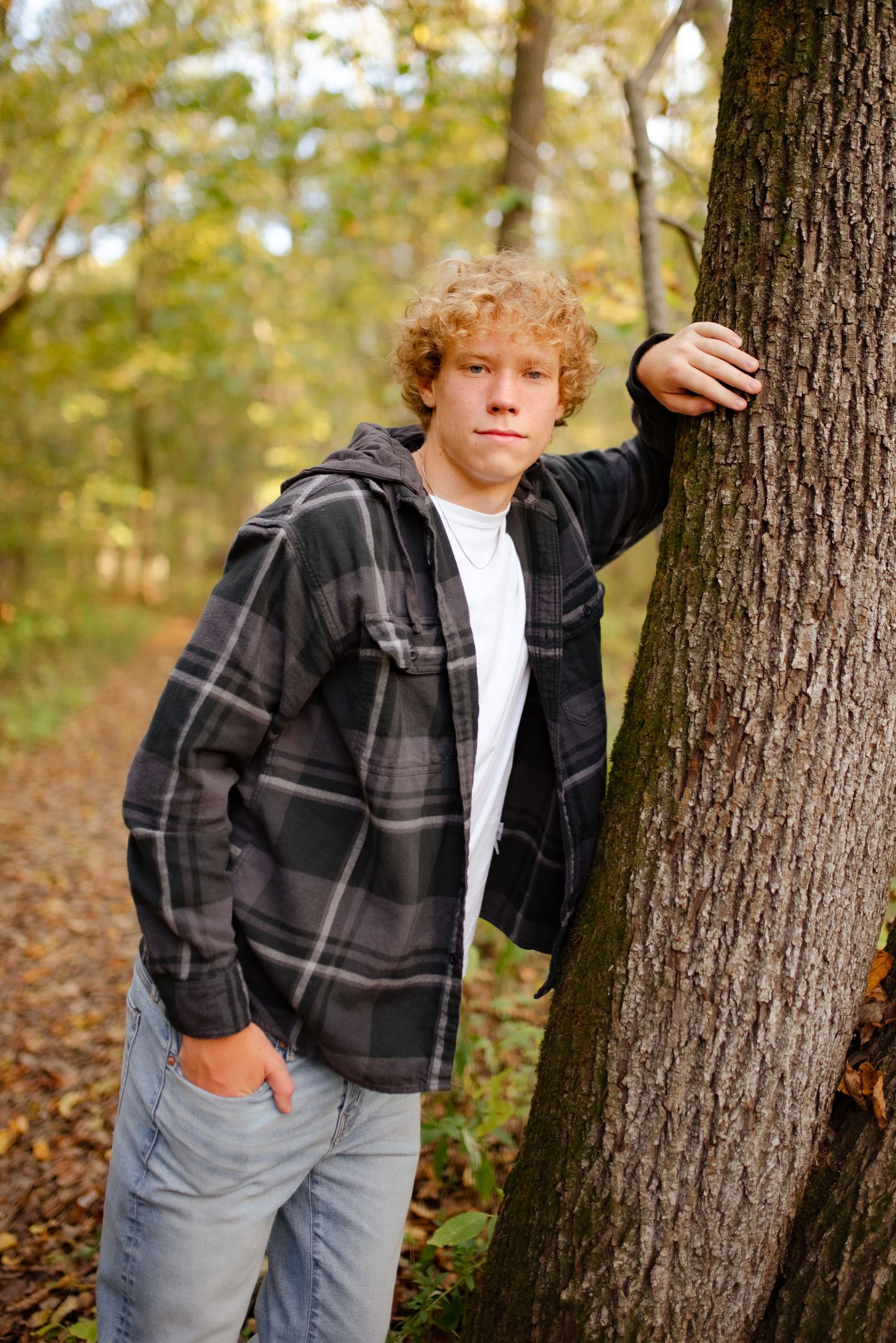 Candid photo of someone leaning against a tree trunk wearing a plaid jacket in a wooded setting.