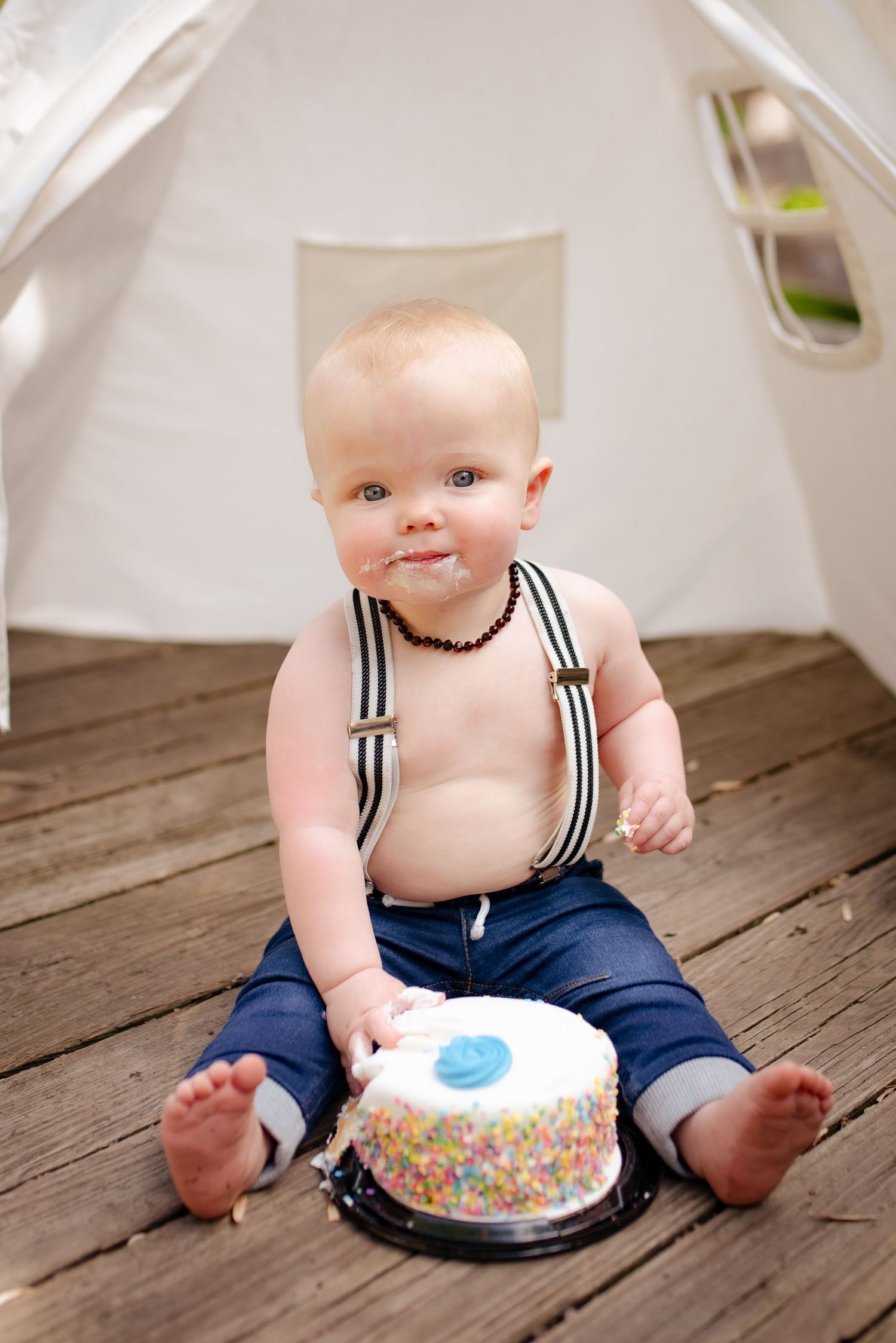 A baby in striped suspenders sits on wooden deck with colorful birthday cake.
