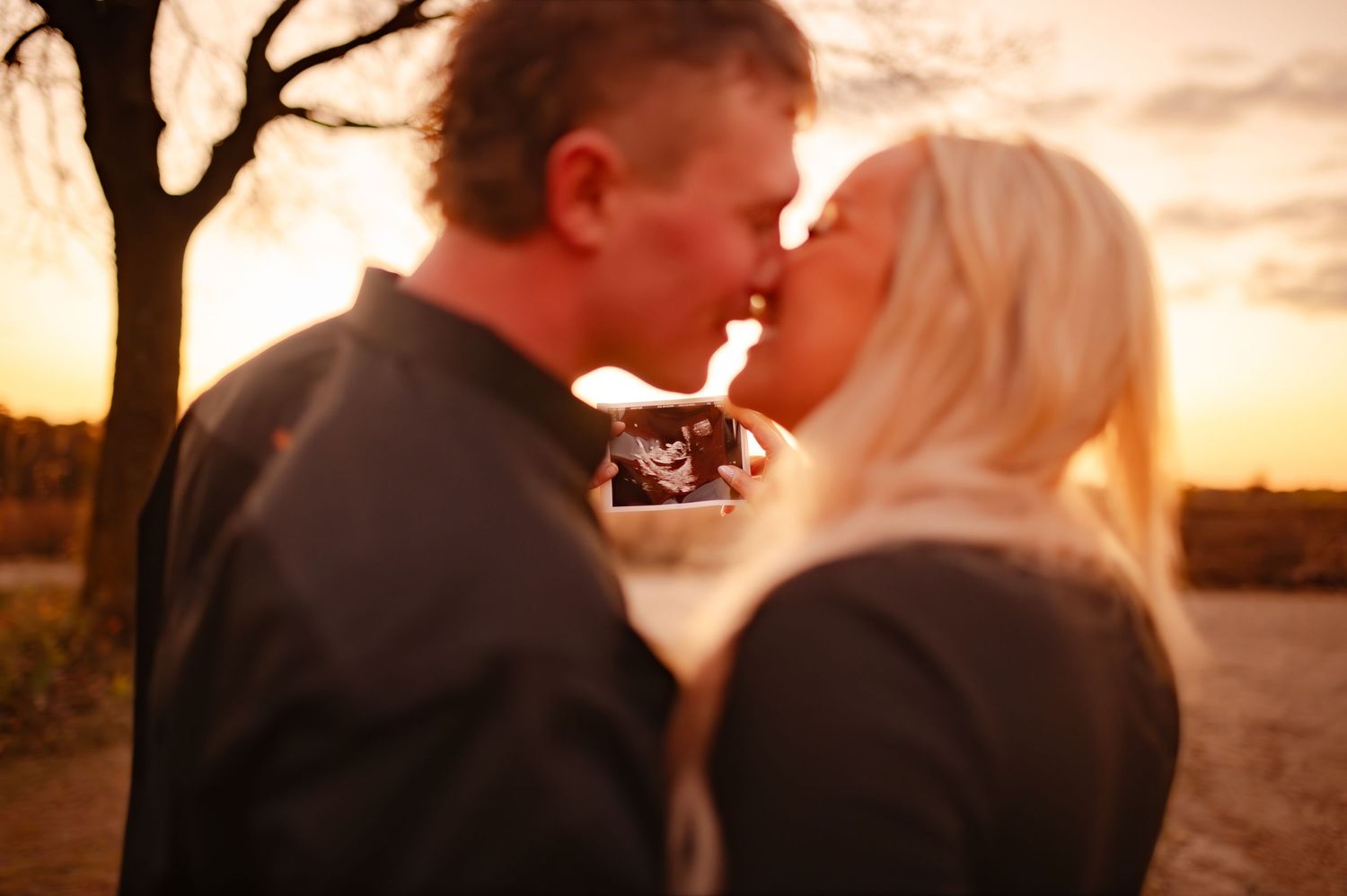 A couple shares a romantic kiss during a golden sunset in a scenic outdoor setting.